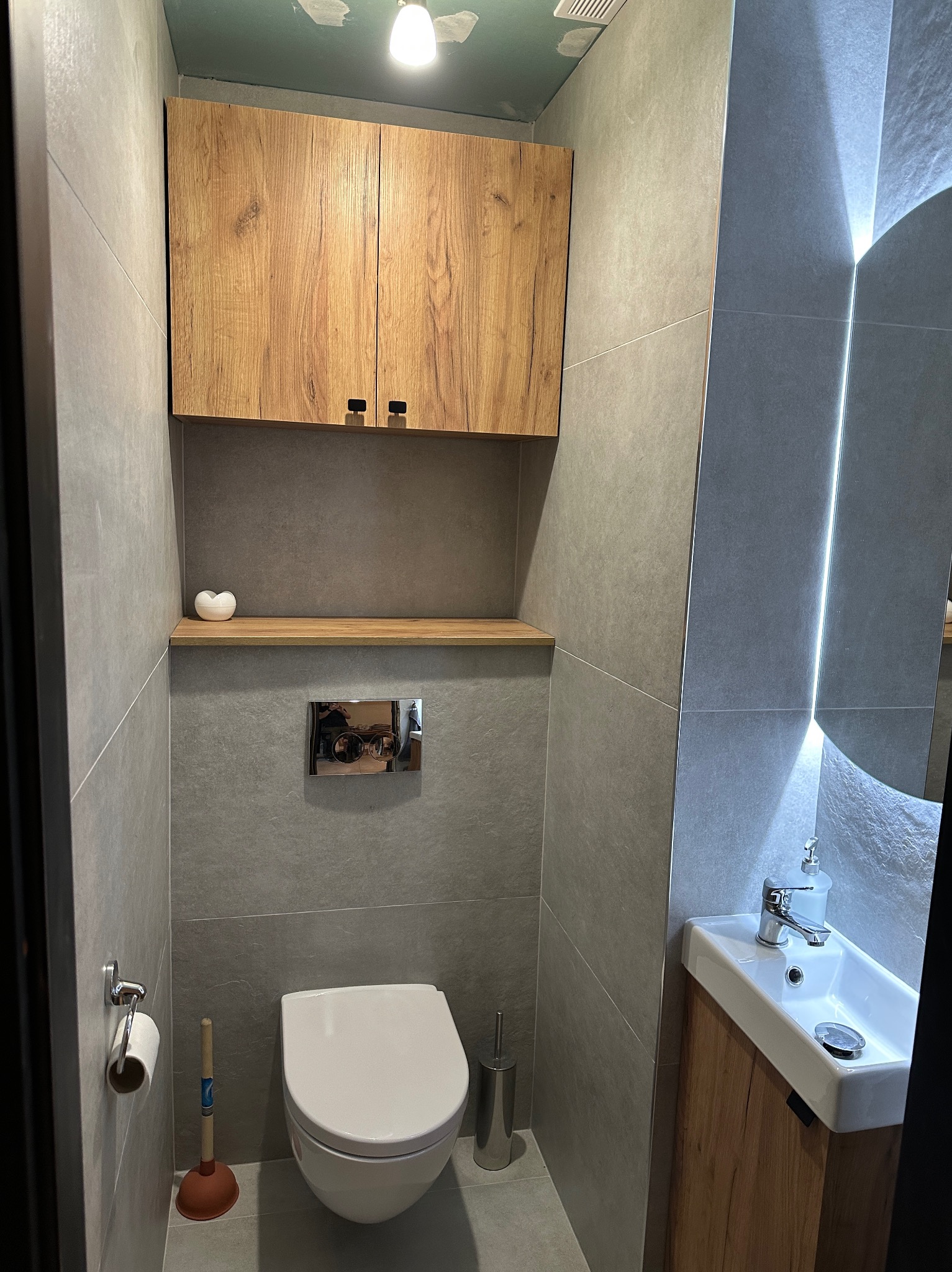 Szara łazienka po remoncie z drewnianymi szafkami, umywalką i toaletą. Na ścianach płytki imitujące kamień, oświetlenie LED w lustrze. Widoczny drobny osprzęt łazienkowy.