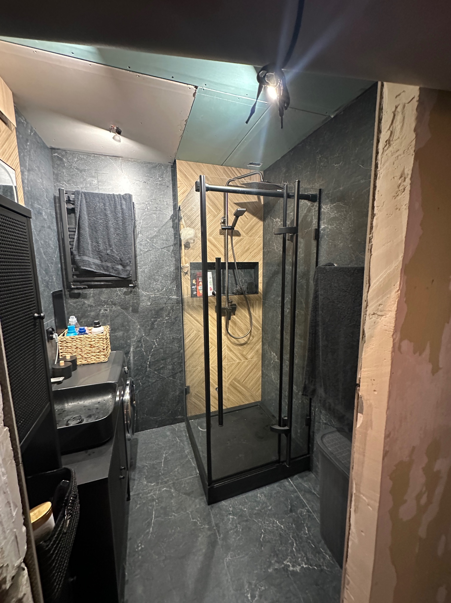 Ciemna łazienka w trakcie remontu: czarna kabina prysznicowa, umywalka i grzejnik na tle marmurowych płytek. Sufit częściowo wykończony, widoczne niedociągnięcia.