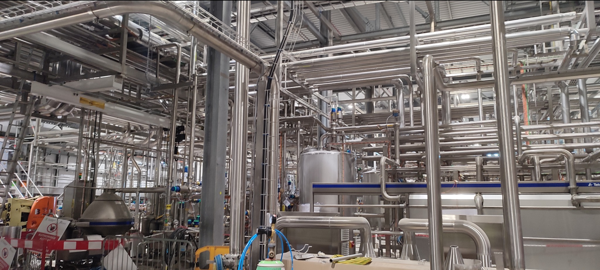 Kompleksowa instalacja rurociągów ze stali nierdzewnej w hali przemysłowej, widoczne liczne złącza i zawory, elementy konstrukcyjne budynku w tle.