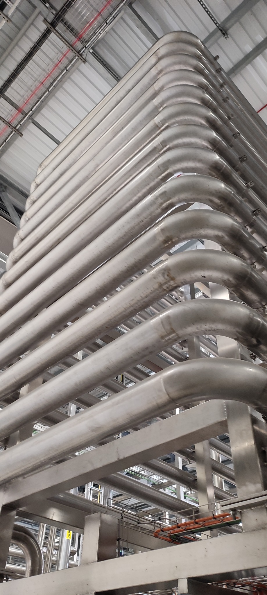 Równoległy układ spawanych rur ze stali nierdzewnej, tworzących zakrzywioną strukturę w przemysłowym otoczeniu, widoczne elementy konstrukcyjne i instalacje pod sufitem.