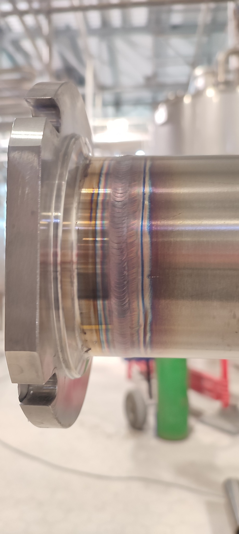 Precyzyjne spawy TIG na rurze ze stali nierdzewnej z kołnierzem, widoczne tęczowe przebarwienia świadczące o wysokiej jakości wykonania. Detal z bliska, w tle warsztat.