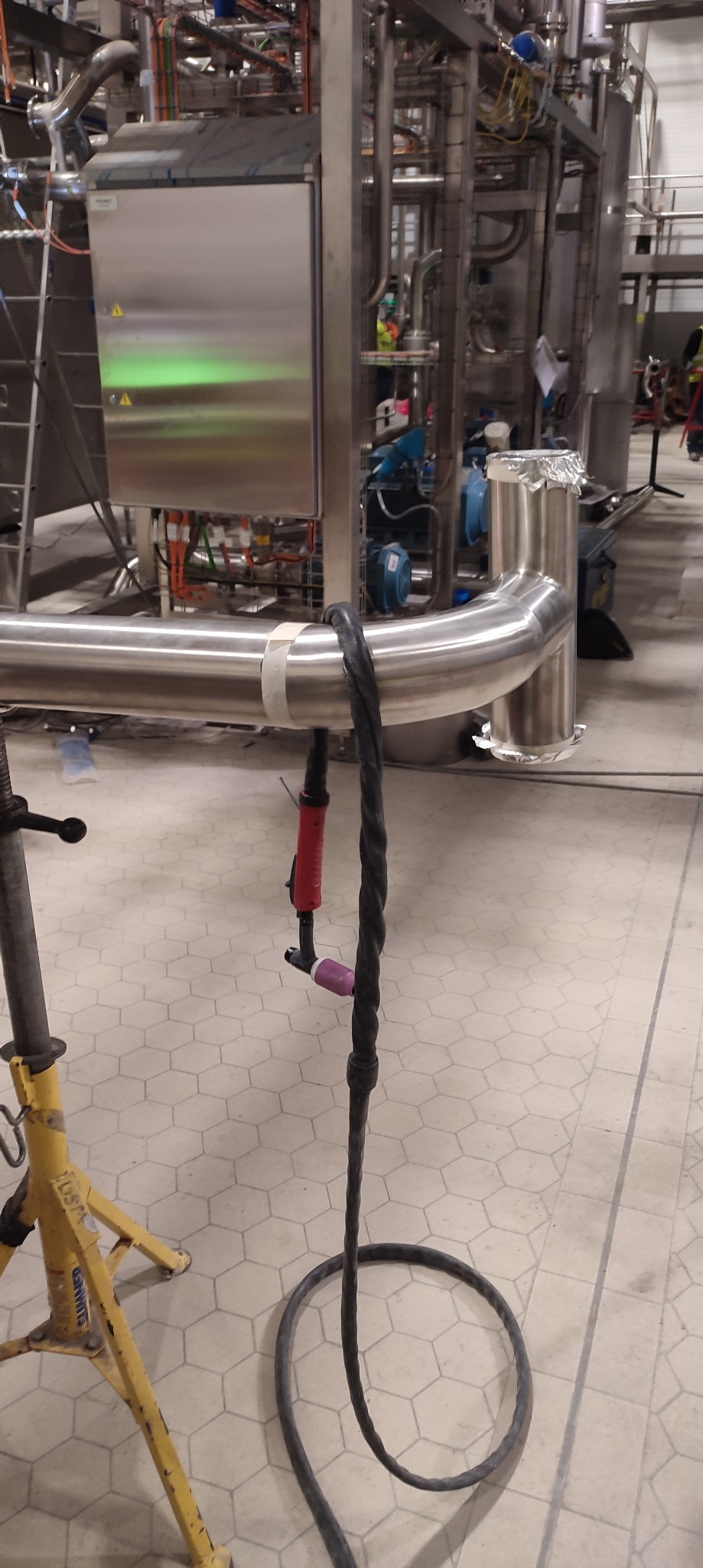 Spoina rury ze stali nierdzewnej w zakładzie przemysłowym, widoczny uchwyt spawalniczy TIG i żółty statyw podtrzymujący rurę. Tło stanowią instalacje przemysłowe.