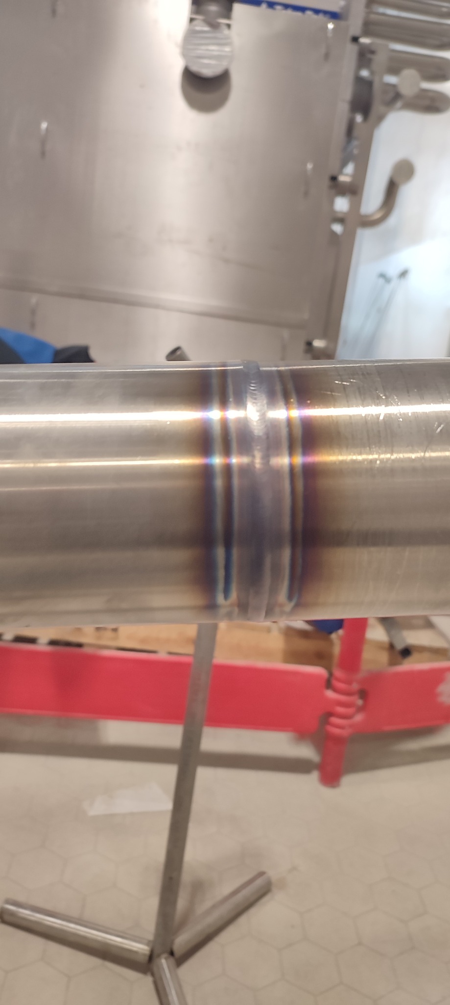 Precyzyjne spoiny na rurze ze stali nierdzewnej, widoczne charakterystyczne przebarwienia po procesie łączenia metali. Tło stanowią elementy wyposażenia warsztatu.