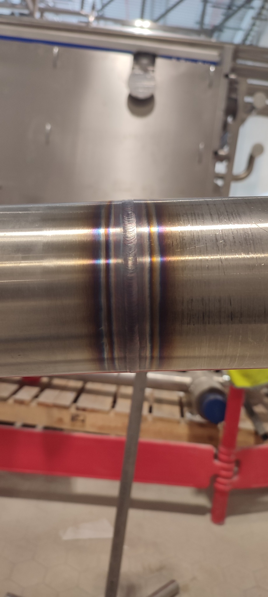 Precyzyjne złącze spawane na rurze ze stali nierdzewnej, widoczne charakterystyczne przebarwienia termiczne. Tło stanowią elementy konstrukcyjne hali.