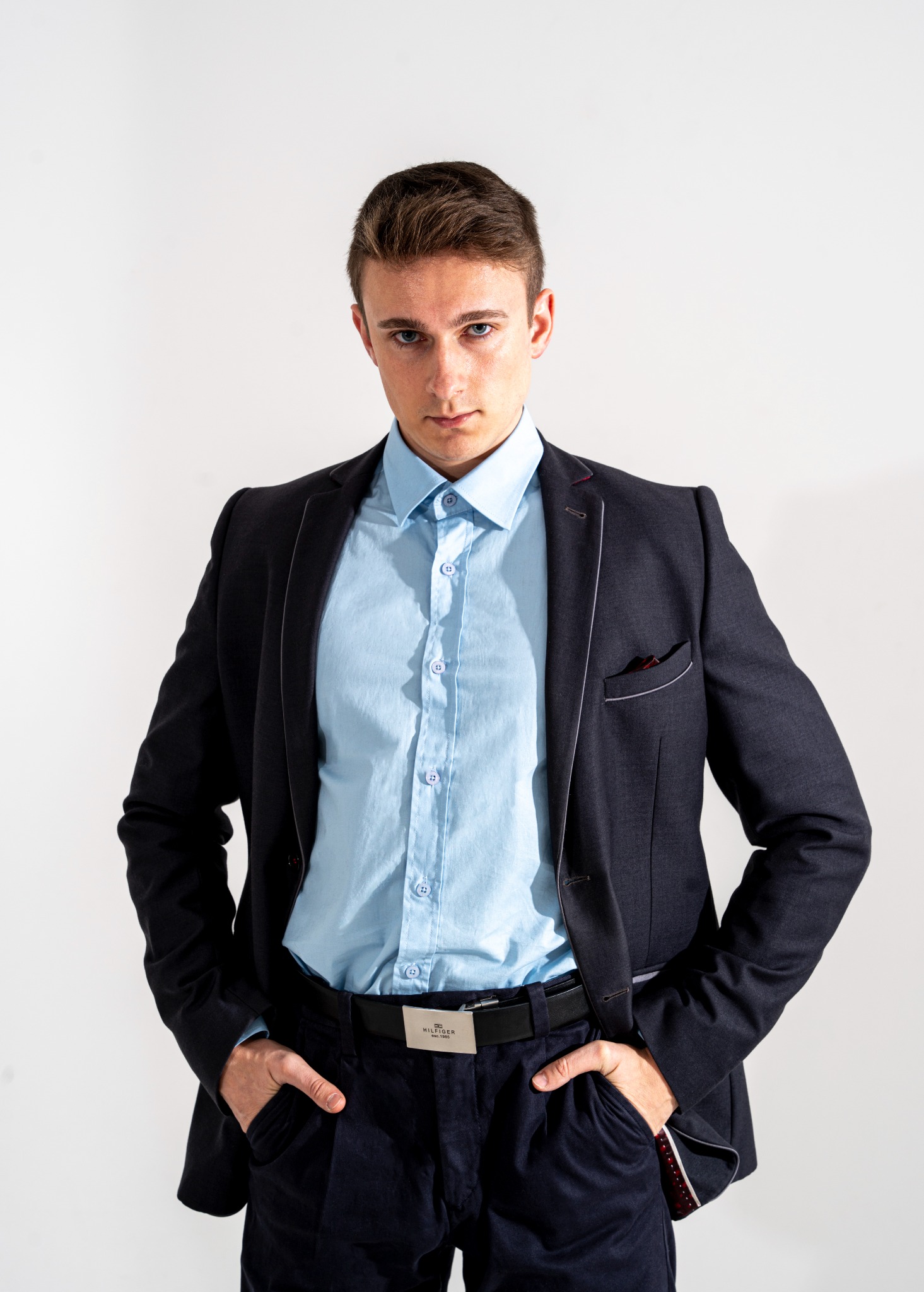 Młody mężczyzna w granatowym garniturze i błękitnej koszuli, pozujący z rękami w kieszeniach na białym tle. Ujęcie od przodu, ostre światło podkreśla rysy twarzy.