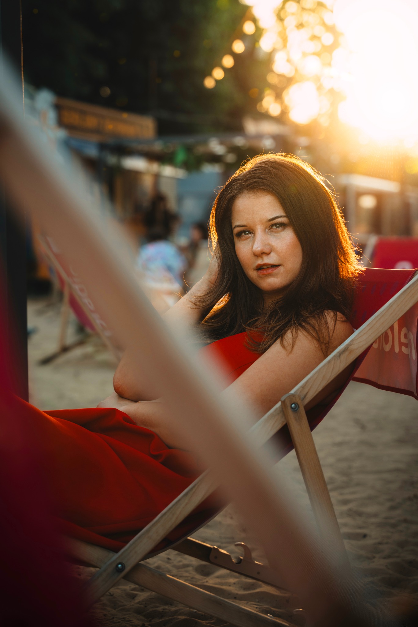 Portret kobiety w czerwonej sukience, siedzącej na leżaku na plaży o zachodzie słońca. Ciepłe światło podkreśla jej włosy i rysy twarzy. W tle widoczne lampki.