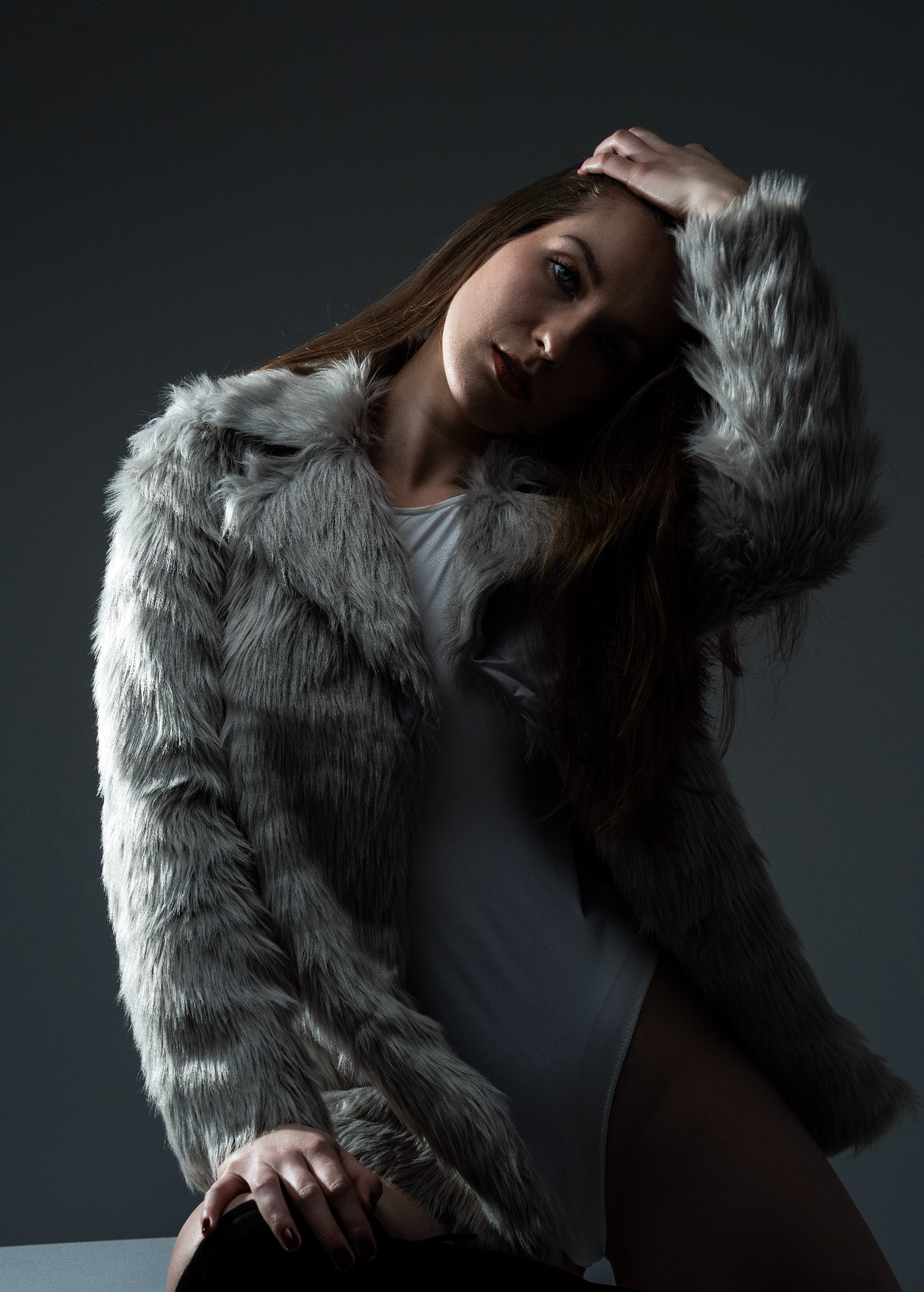 Portret kobiety w futrzanym płaszczu na szarym tle. Modelka pozuje z ręką na głowie, spoglądając w obiektyw. Ujęcie w klimacie fashion z subtelnym oświetleniem.