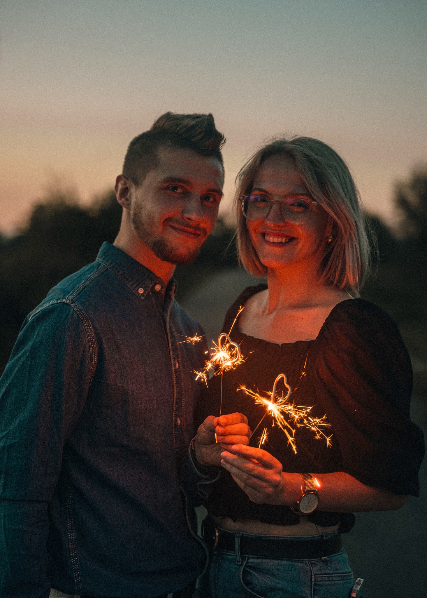 Uśmiechnięta para trzymająca zimne ognie w Nowym Brzesku, na tle zachodzącego słońca. Kobieta w okularach i zegarku, mężczyzna w jeansowej koszuli. Romantyczna atmosfera.