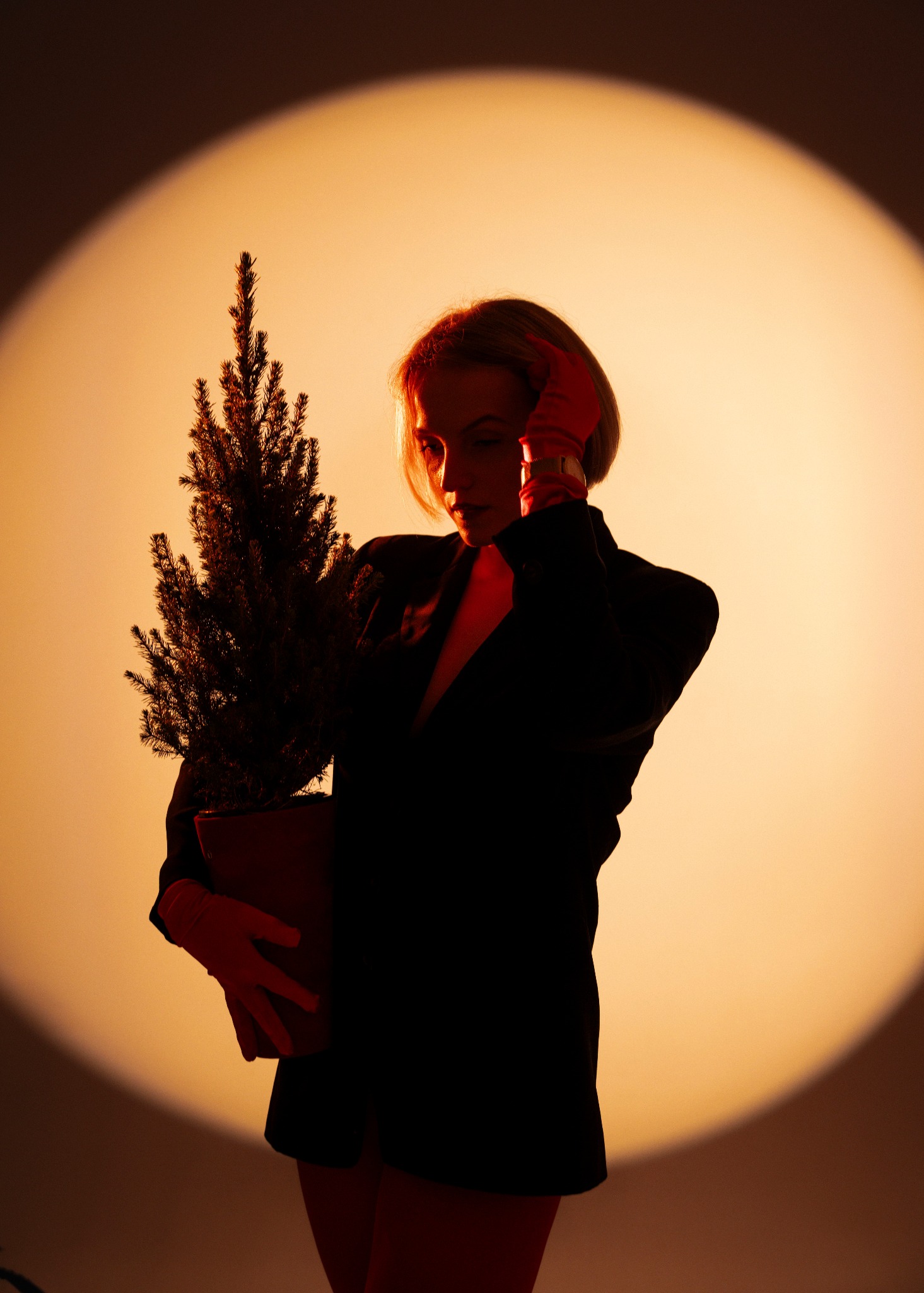 Kobieta w czarnej marynarce i czerwonych rękawiczkach trzyma małą choinkę na pomarańczowym tle z efektem reflektora. Nietypowe ujęcie, artystyczna fotografia.