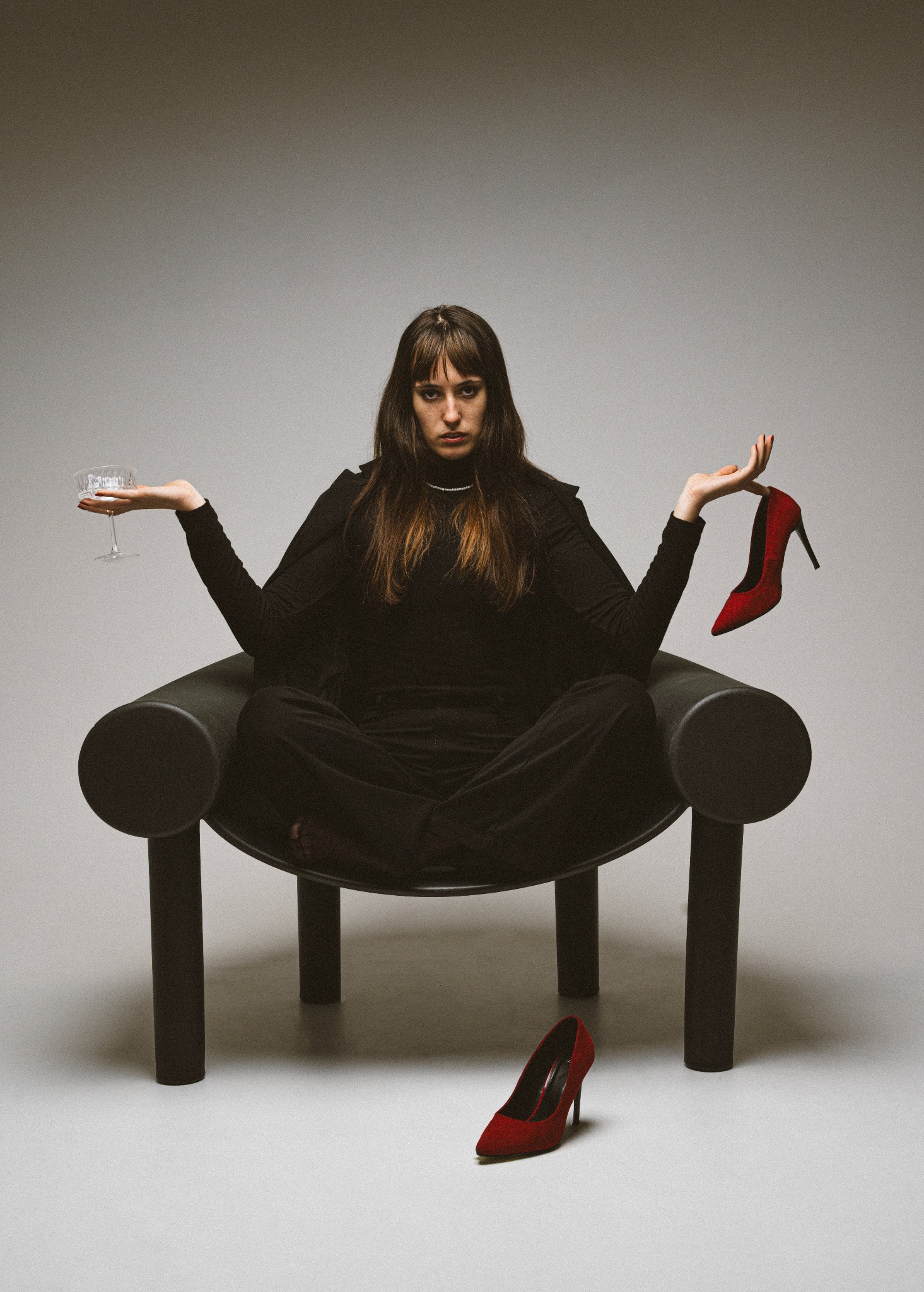 Kobieta w czarnym stroju siedzi na krześle, trzymając kieliszek i czerwony szpilkę. Druga szpilka leży na podłodze. Minimalistyczne tło, centralna kompozycja.