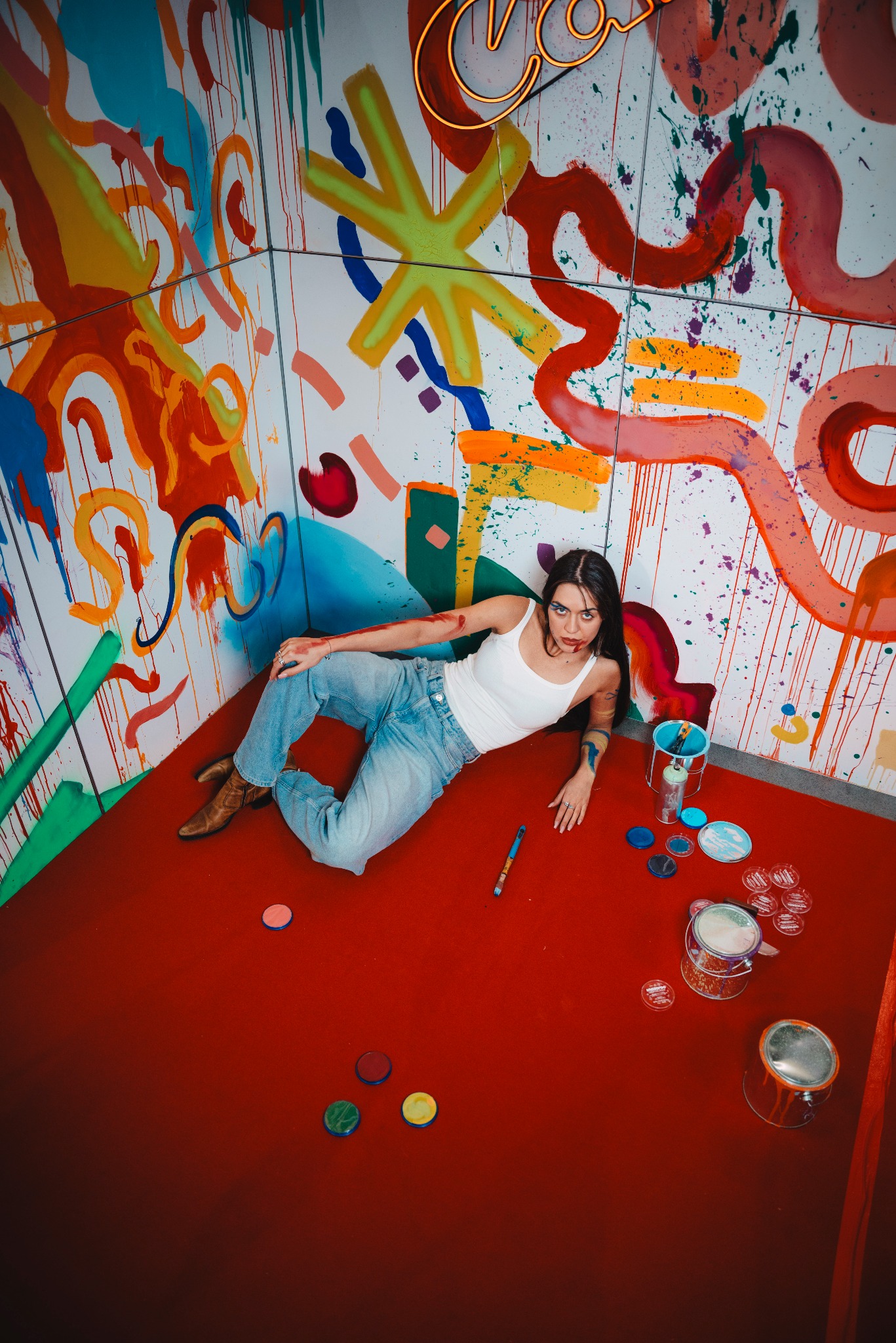 Młoda kobieta leży na czerwonej podłodze w pokoju z abstrakcyjnymi malowidłami na ścianach, otoczona farbami i pędzlami, spoglądając w górę.