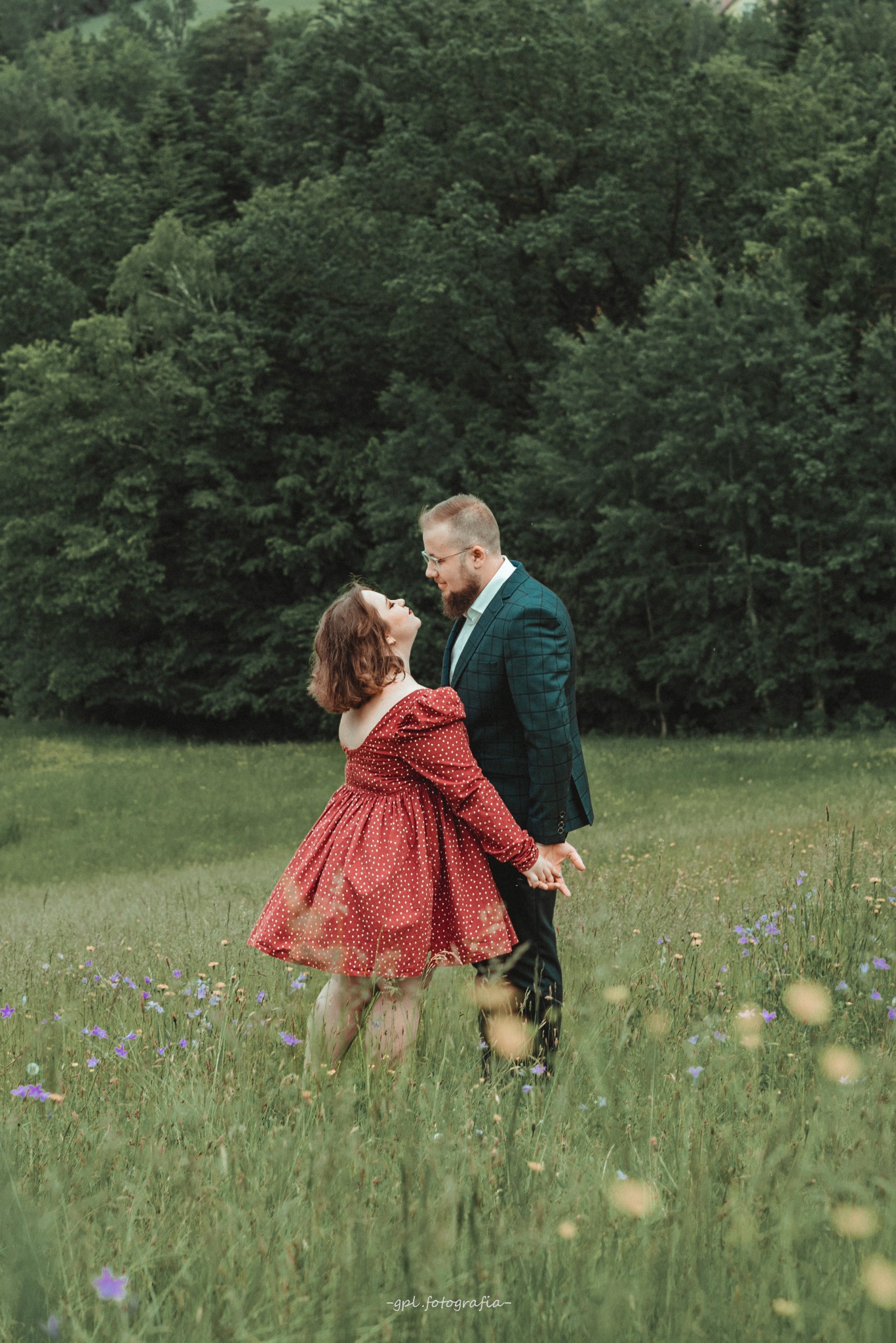 Zakochana para w plenerze: kobieta w czerwonej sukience i mężczyzna w garniturze, trzymają się za ręce na łące z kwiatami, zalesione wzgórze w tle.