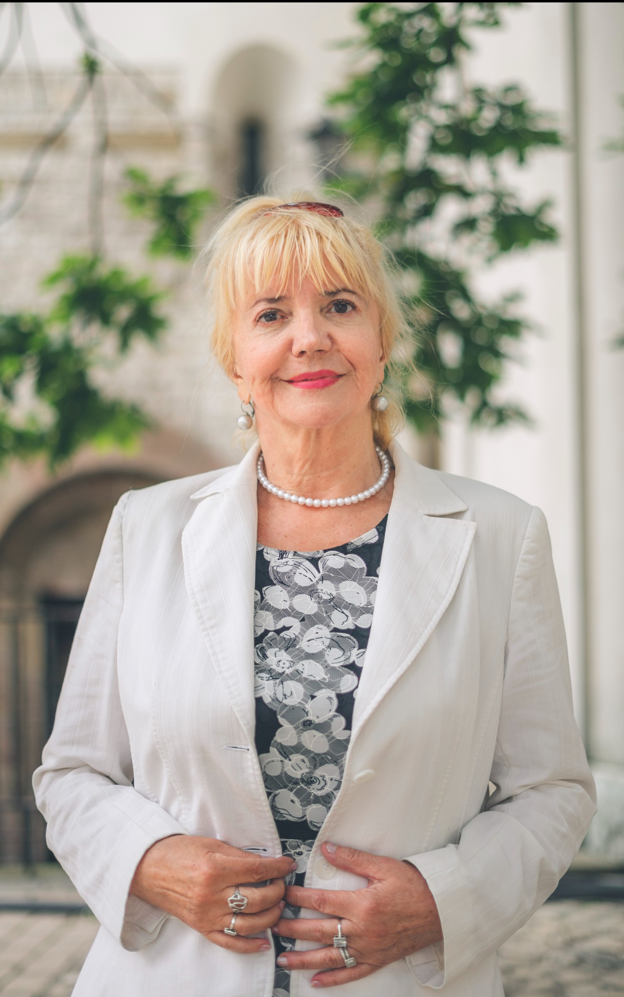 Portret uśmiechniętej kobiety w białym żakiecie, z perłami i pierścionkami, na tle zieleni i jasnej architektury. Ujęcie z delikatnym rozmyciem tła.