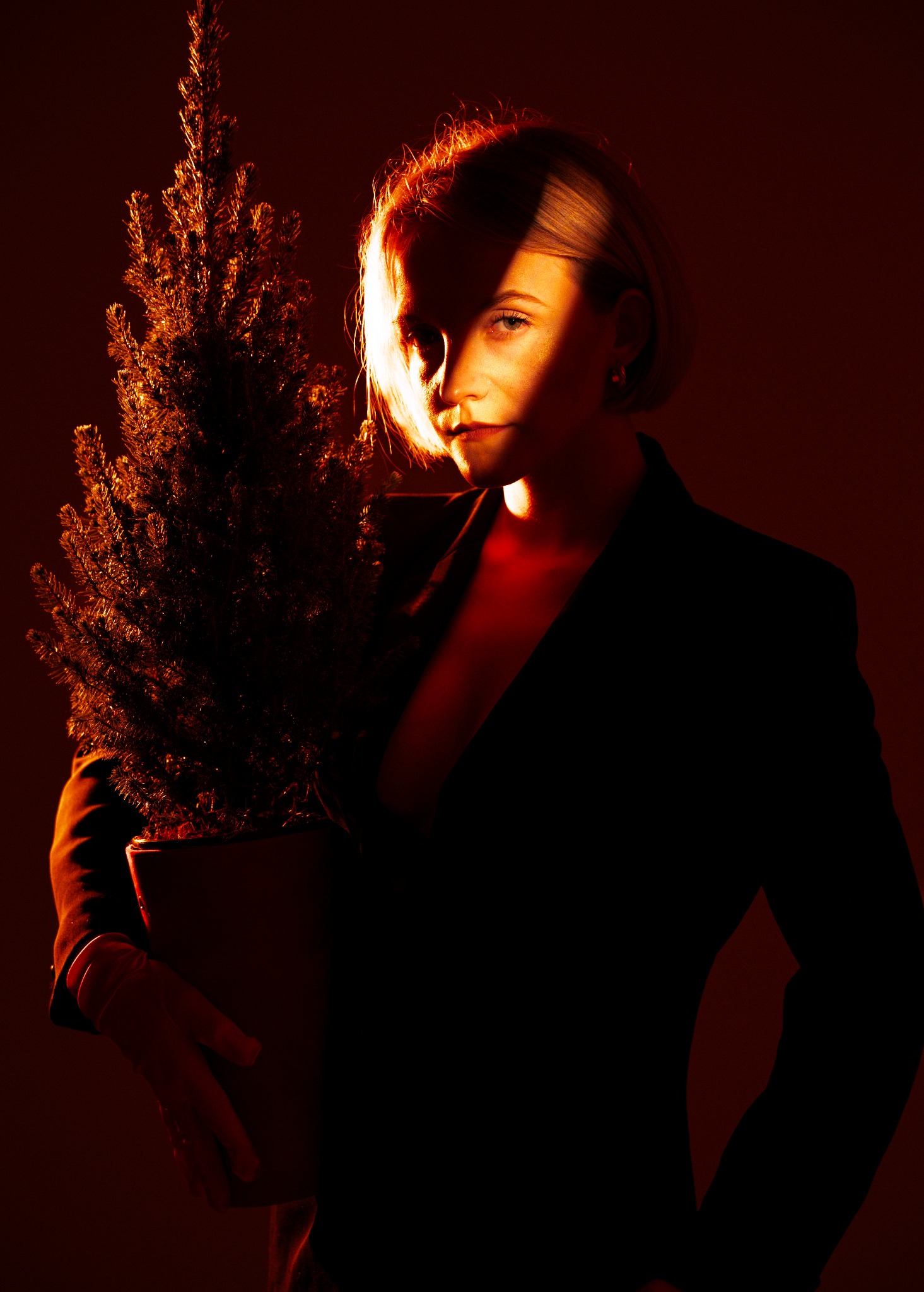 Portret kobiety w marynarce, trzymającej małą choinkę w doniczce, oświetlonej ciepłym, pomarańczowym światłem, tworzącym dramatyczny cień na twarzy.