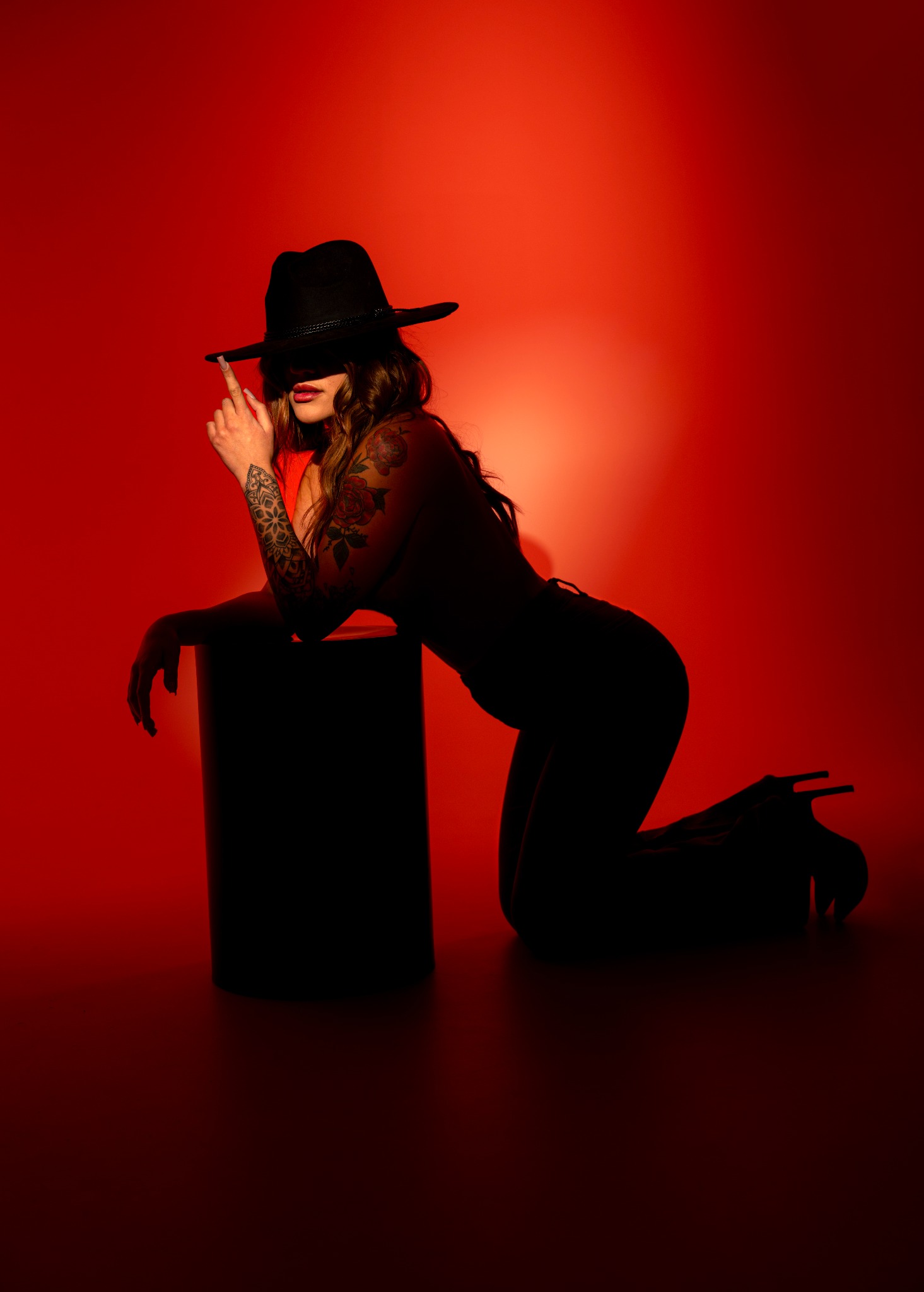 Kobieta w kapeluszu, z tatuażami, pozuje na kolanach, opierając się o czarny cylinder na intensywnie czerwonym tle. Oświetlenie tworzy dramatyczny efekt cieni.