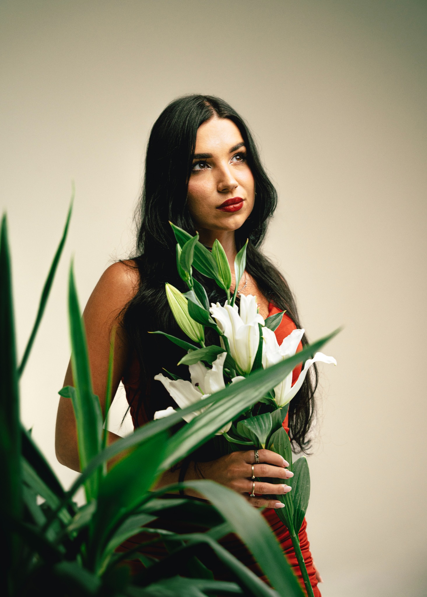 Portret kobiety z ciemnymi włosami i czerwonymi ustami, trzymającej bukiet białych lilii, z zieloną rośliną w pierwszym planie, spoglądającej w górę.