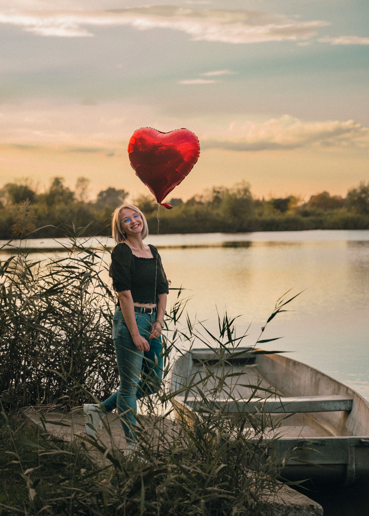 Uśmiechnięta blondynka w dżinsach i czarnej bluzce, trzymająca czerwony balon w kształcie serca nad jeziorem, obok łódki i trzcin, w ciepłym świetle zachodzącego słońca.