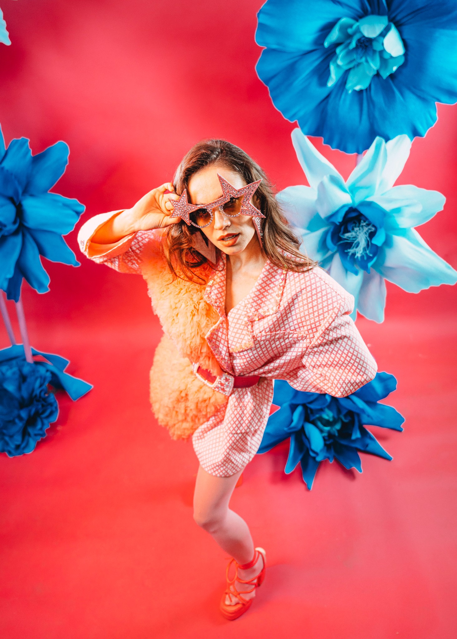Kobieta w różowej sukience i okularach w kształcie gwiazd pozuje na tle czerwonego studia z niebieskimi kwiatami. Nietypowa sesja w stylu retro.