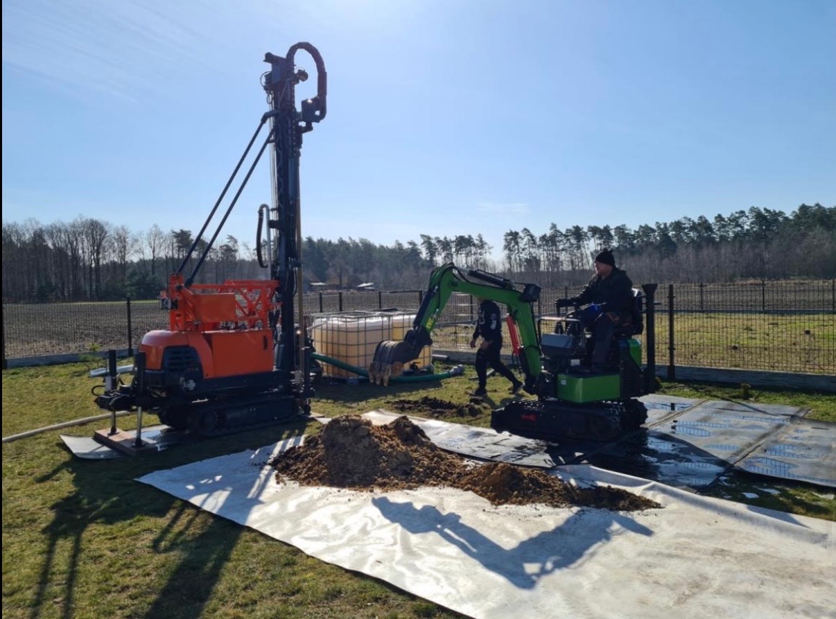 Wiertnica do studni głębinowych i minikoparka na trawniku, zabezpieczonym folią. W tle drzewa i ogrodzenie z siatki. Błękitne niebo.