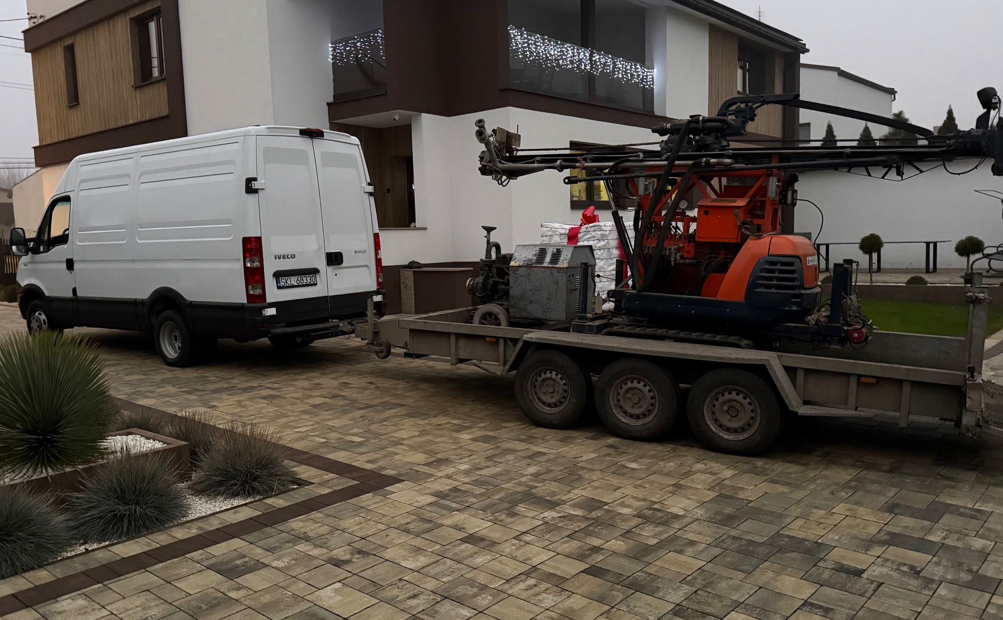 Biały bus Iveco z przyczepą i wiertnicą na posesji z kostki brukowej. W tle dom z oświetleniem.