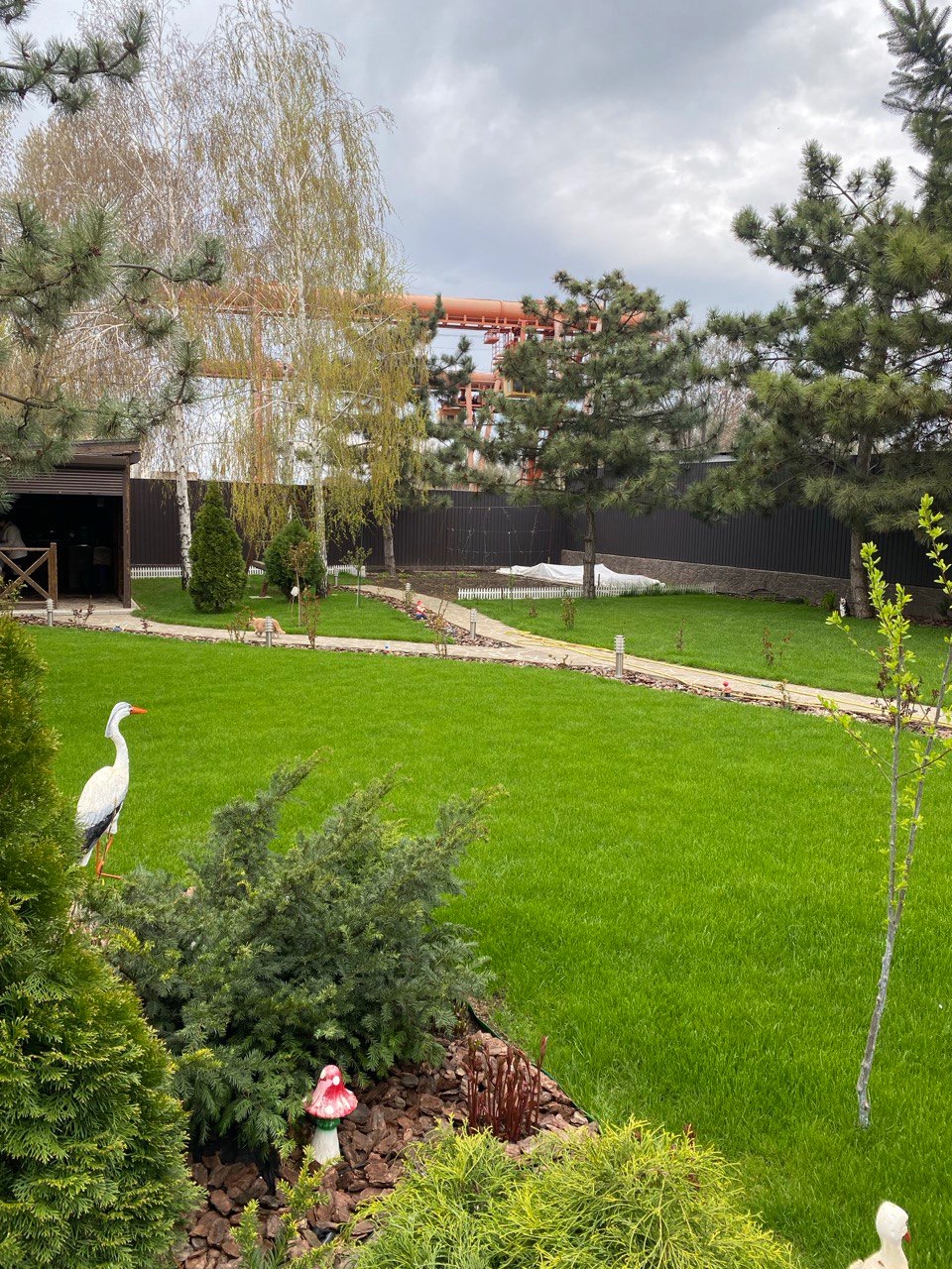 Zadbany trawnik z dekoracjami ogrodowymi: figurka bociana, muchomorek, krzewy ozdobne. W tle ścieżka, altana i drzewa iglaste. Całość w otoczeniu ogrodzenia.