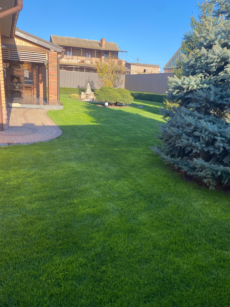 Zadbany trawnik z elementami architektury ogrodowej, widok na dom i ogrodzenie. Świeżo skoszona trawa, dekoracyjne krzewy i drzewa iglaste.