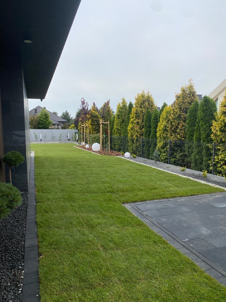 Nowoczesny ogród z równo przystrzyżonym trawnikiem, czarną kostką brukową i ozdobnymi kulami świetlnymi wzdłuż ogrodzenia z tujami. Minimalistyczny design.