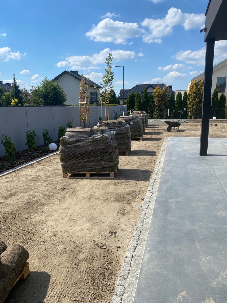 Przygotowane rolki trawy na paletach, czekające na rozłożenie na wyrównanym terenie. Widoczne obrzeża i fragment tarasu. Słoneczny dzień.