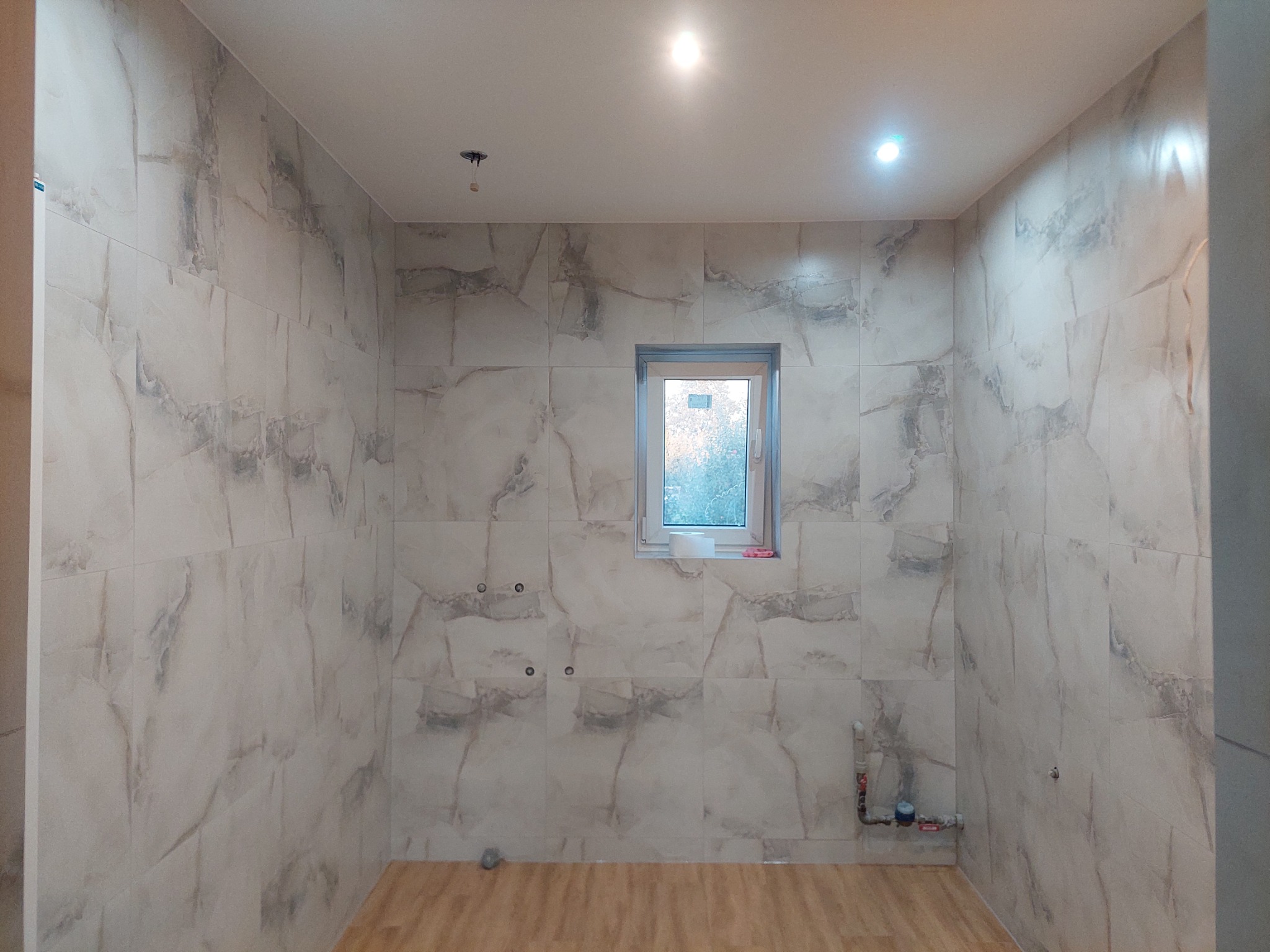 Wykończona łazienka z marmurowymi płytkami, drewnopodobną podłogą i oknem. Widoczne punkty instalacji wodnej i oświetlenie sufitowe.