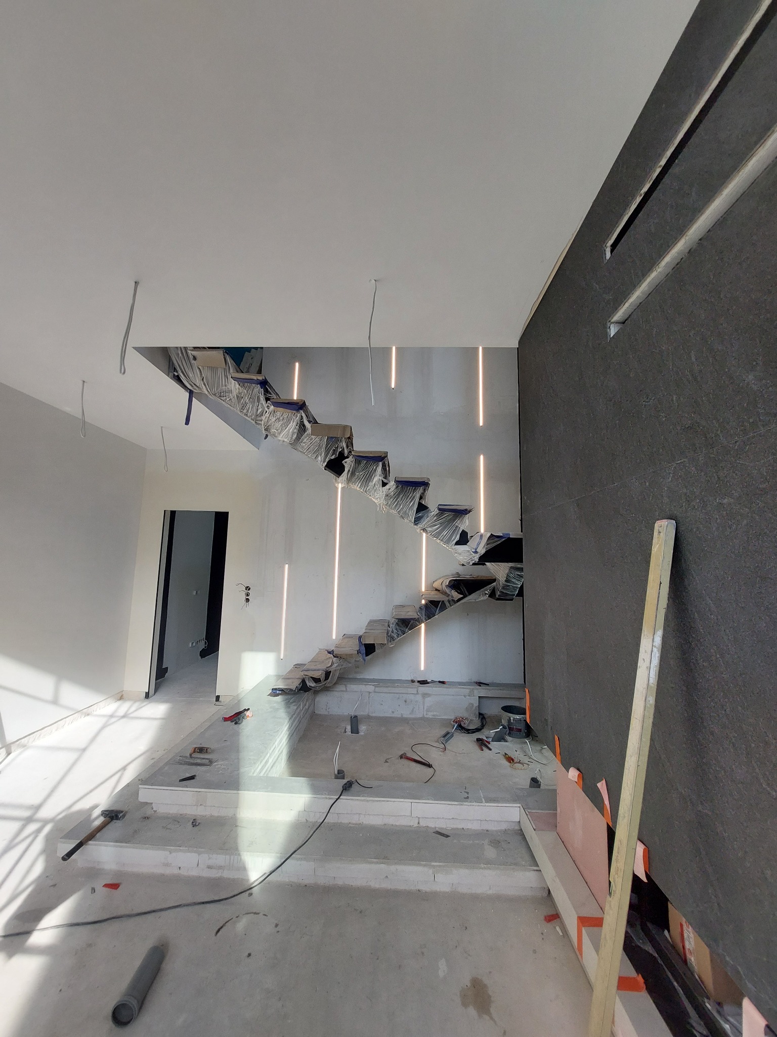 Wnętrze w trakcie wykończenia: schody z podświetleniem, ściana z ciemnej płyty, widoczne narzędzia i materiały budowlane. Etap prac wykończeniowych w nowym domu.