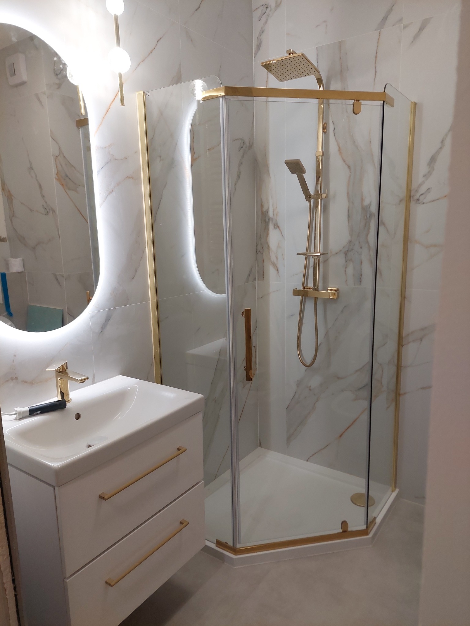 Nowoczesna łazienka z kabiną prysznicową w złotych ramach i umywalką z szafką. Marmurowe płytki na ścianach i podświetlane lustro owalne dodają elegancji.
