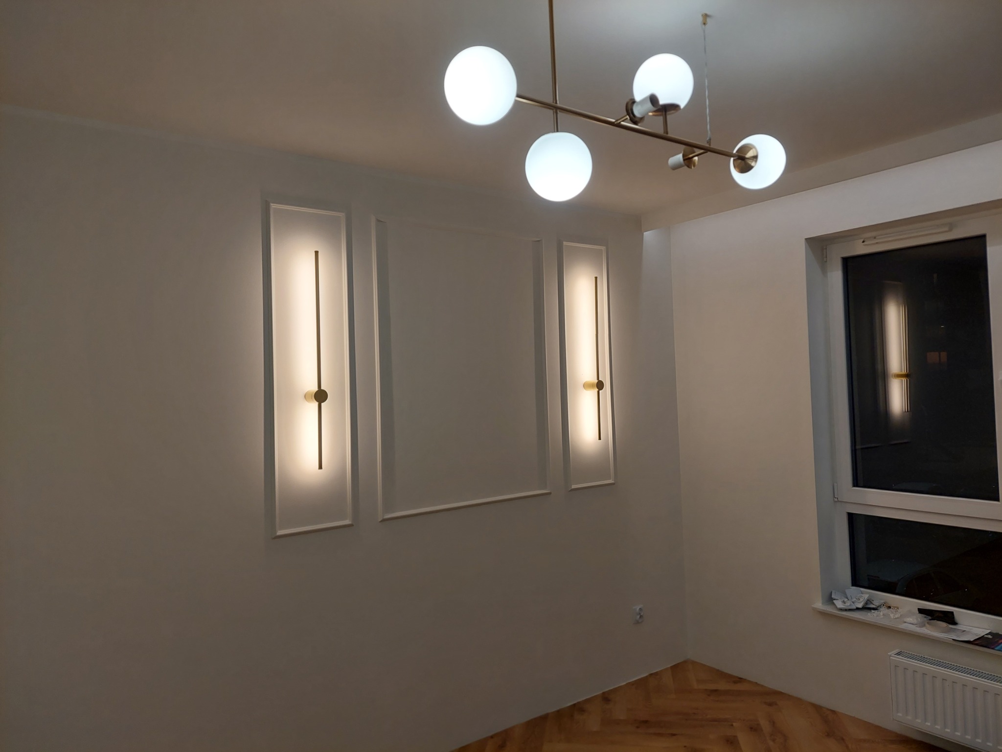 Białe ściany z dekoracyjnymi, podświetlanymi panelami w ramkach i nowoczesną lampą sufitową z kulistymi kloszami w wyremontowanym pokoju z parkietem i oknem.