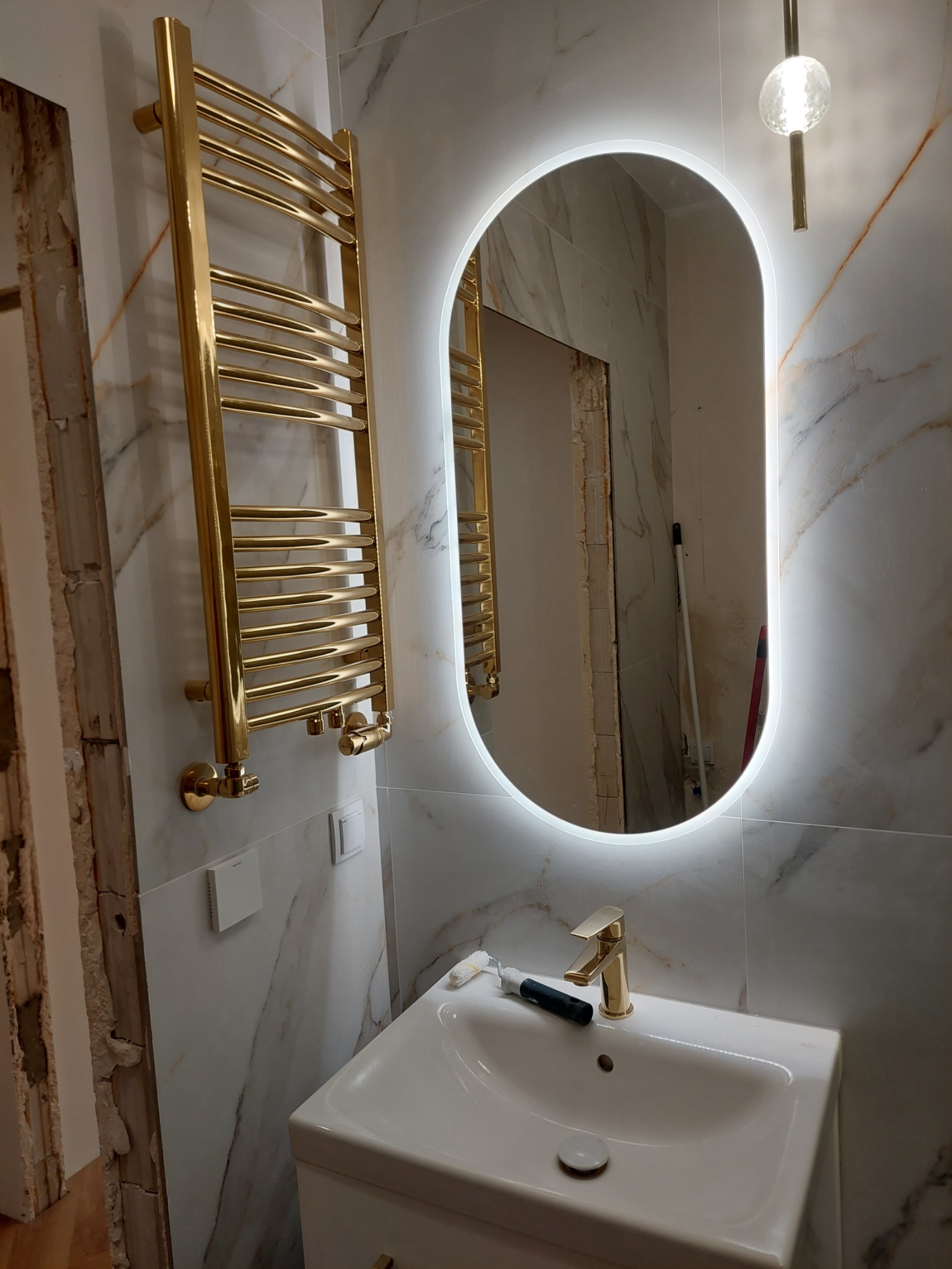 Wykończona łazienka z owalnym lustrem LED, złotą armaturą i grzejnikiem drabinkowym. Marmurowe płytki na ścianach. Widoczny fragment niezakończonego wykończenia obok.