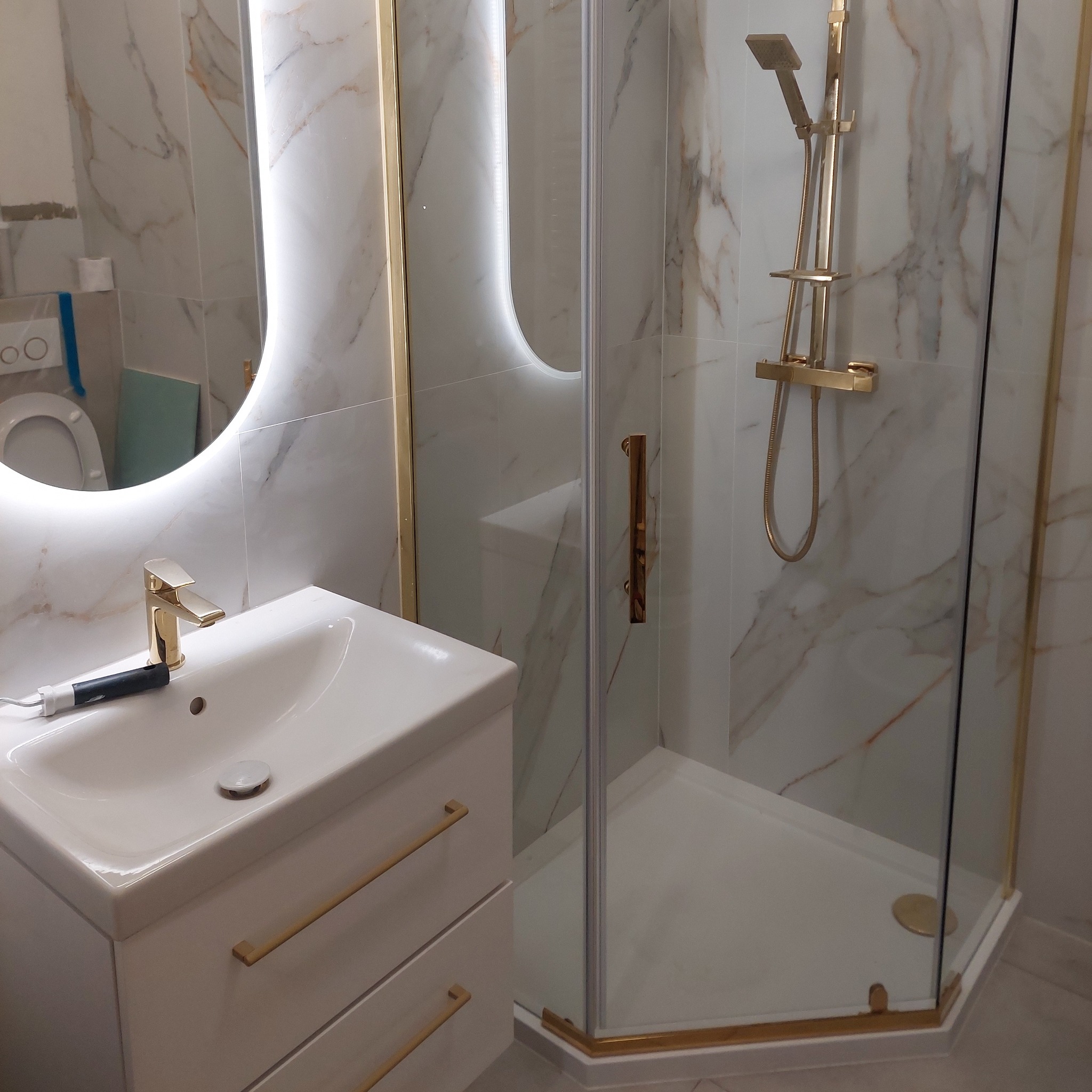 Wykończona łazienka z kabiną prysznicową i umywalką w stylu glamour. Złote akcenty na armaturze i obramowaniach, marmurowe płytki na ścianach, oświetlone lustro.