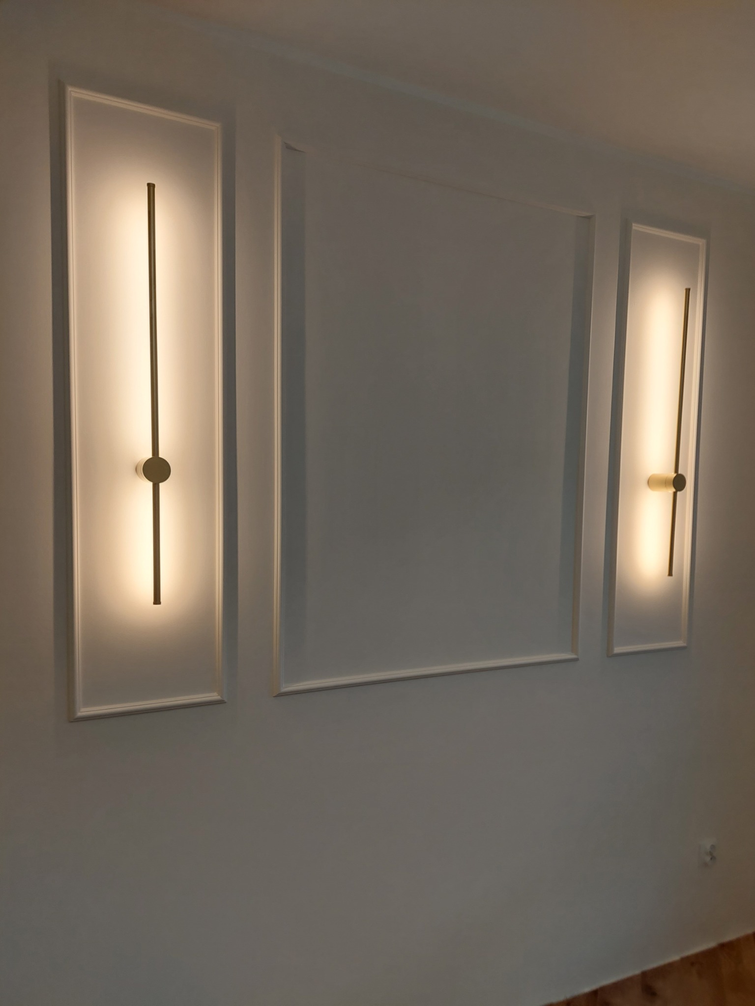 Ściana z dekoracyjnymi, białymi panelami i dwoma podłużnymi lampami LED z elementami w kolorze złota, oświetlającymi ścianę. Minimalistyczny design.