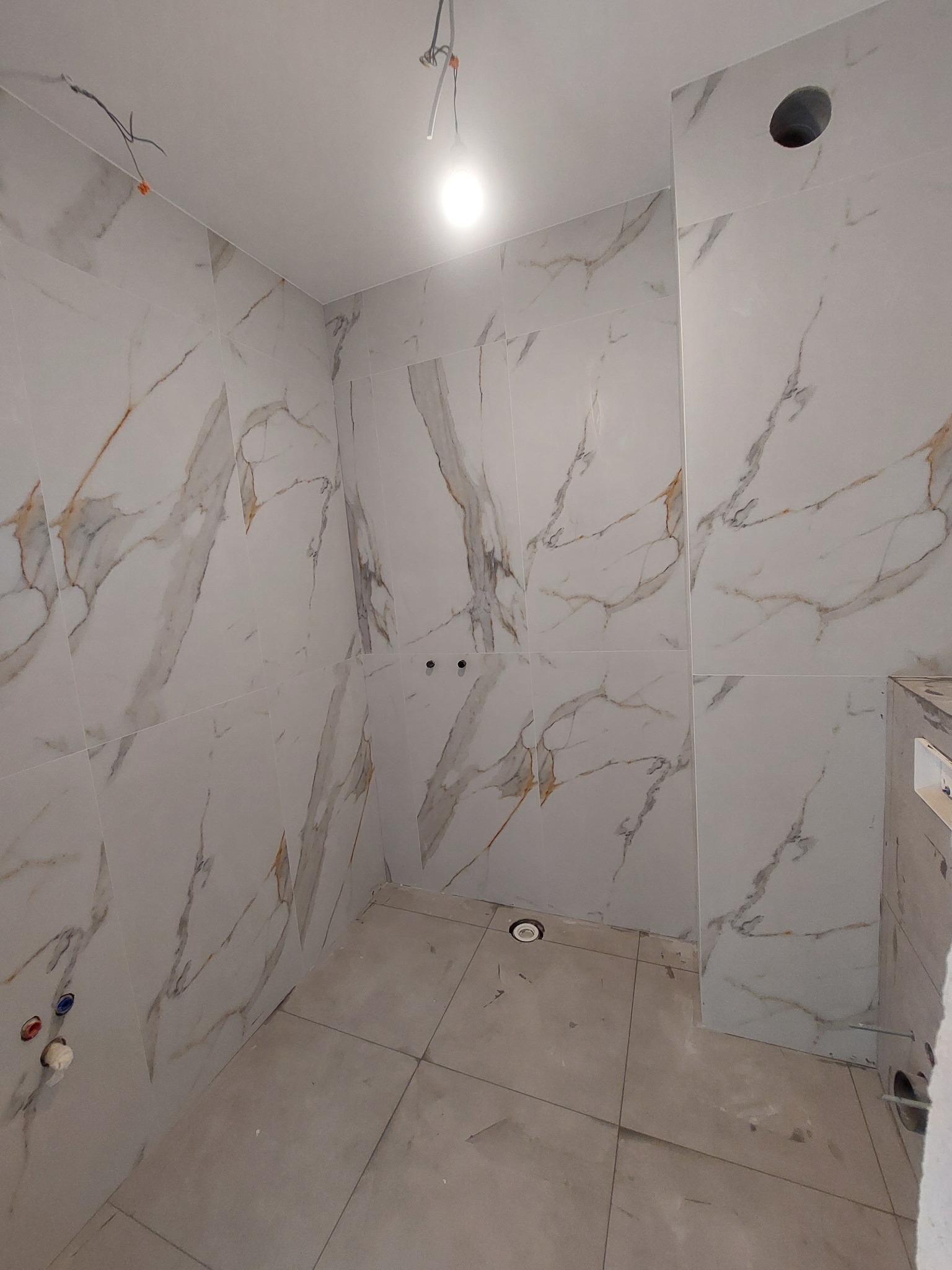 Wykończona łazienka z marmurowymi płytkami na ścianach i podłodze. Widoczne otwory na armaturę i oświetlenie. Minimalistyczny design z elementami instalacji.