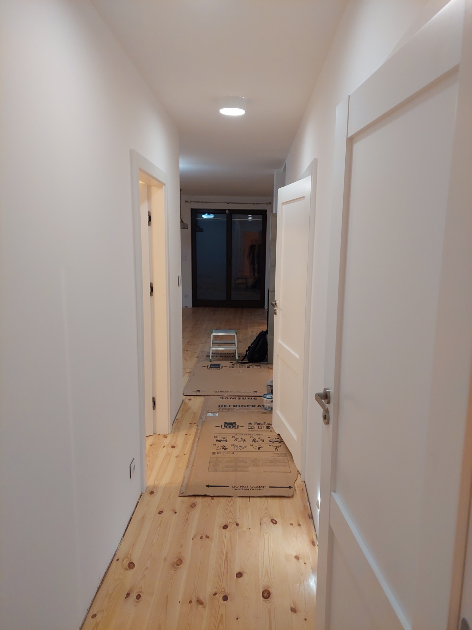 Jasny korytarz z drewnianą podłogą zabezpieczoną kartonami, białe ściany i drzwi. Widoczna drabina i torba w głębi. Przygotowanie do remontu lub po ułożeniu podłogi.
