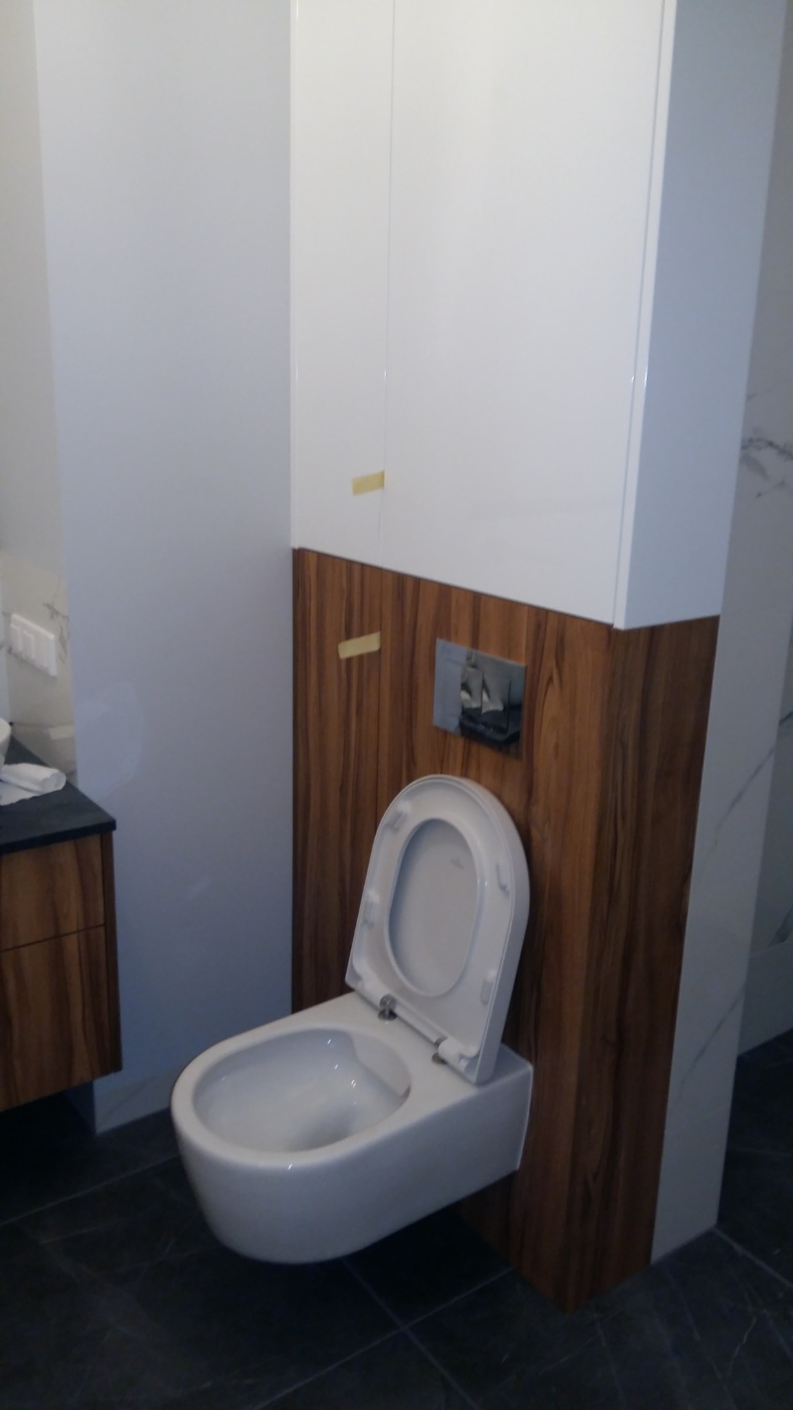 Nowoczesna toaleta podwieszana z deską otwartą, obudowana drewnopodobną płytą, wkomponowana w białą zabudowę łazienkową. Widoczne płytki podłogowe i fragment szafki.