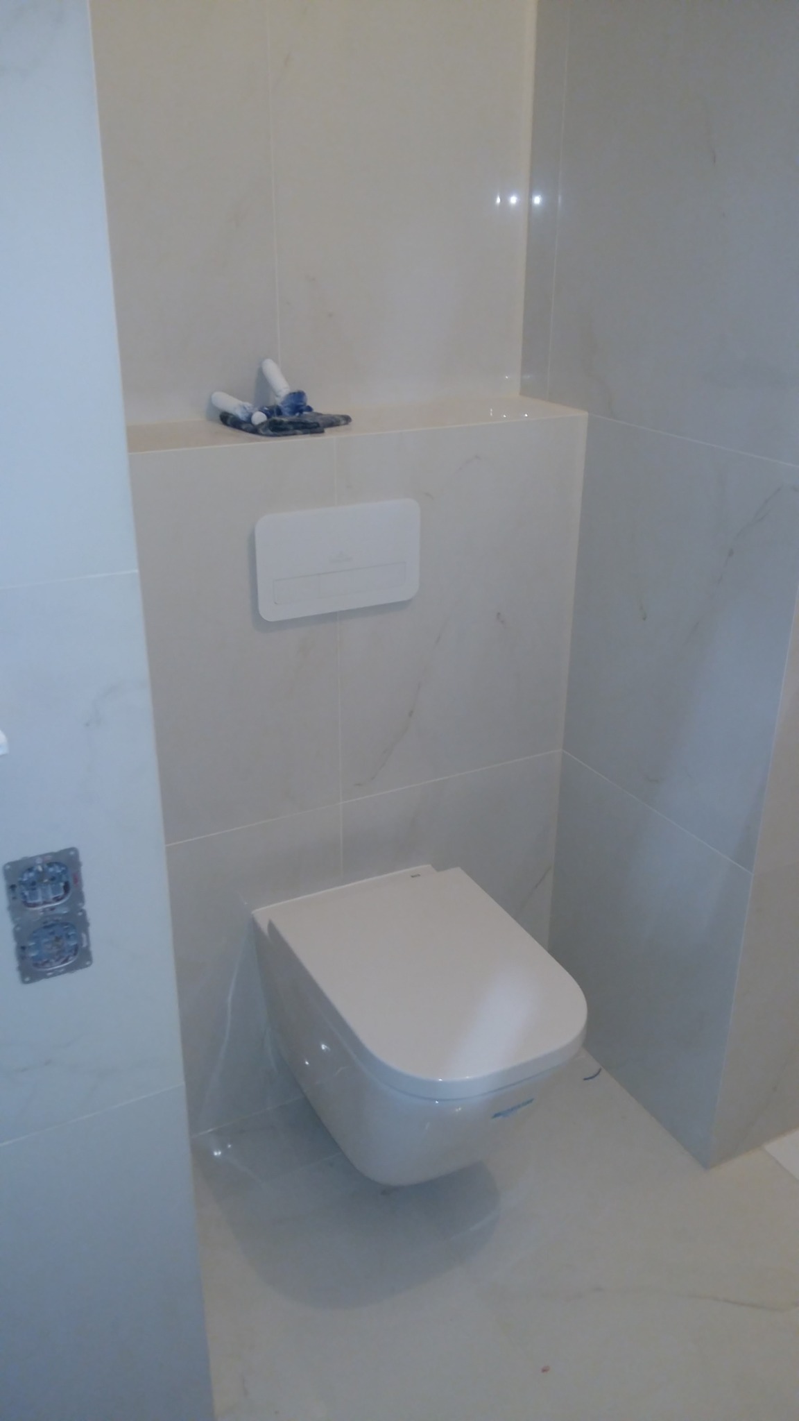 Elegancka, podwieszana toaleta w wykończonej łazience, z płytkami imitującymi marmur i ukrytym systemem spłukiwania. Widoczne gniazdka elektryczne po lewej stronie.