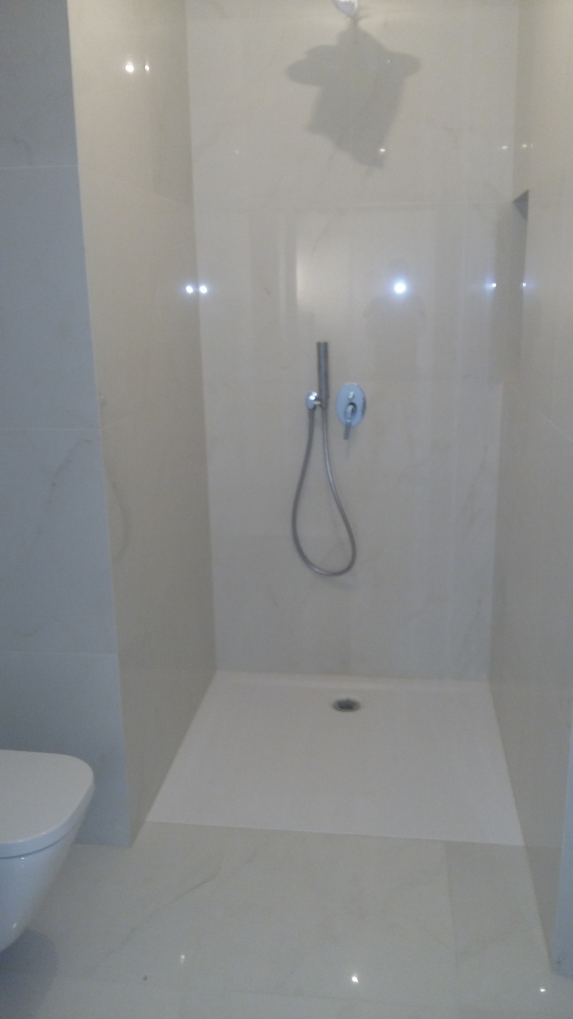 Jasna, wykafelkowana kabina prysznicowa z deszczownicą i baterią, widoczny fragment toalety. Minimalistyczny design, marmurowe płytki, czysta przestrzeń.