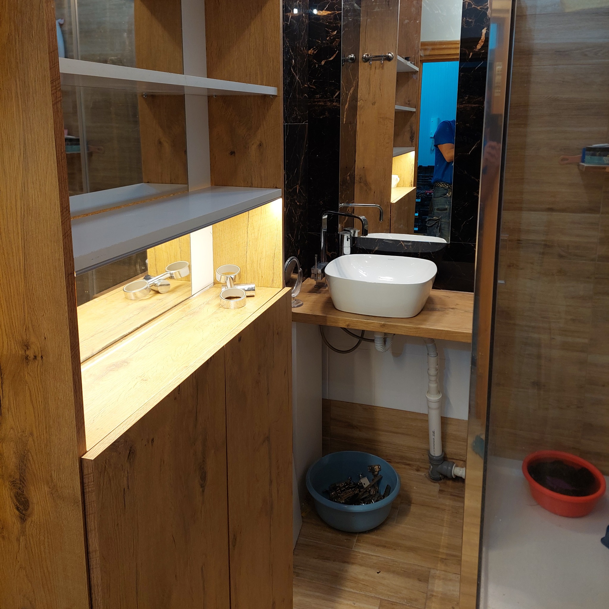 Nowoczesna łazienka z drewnianymi szafkami, białą umywalką nablatową i czarną ścianą z marmurowym wzorem. Widoczny fragment lustra i akcesoria łazienkowe.