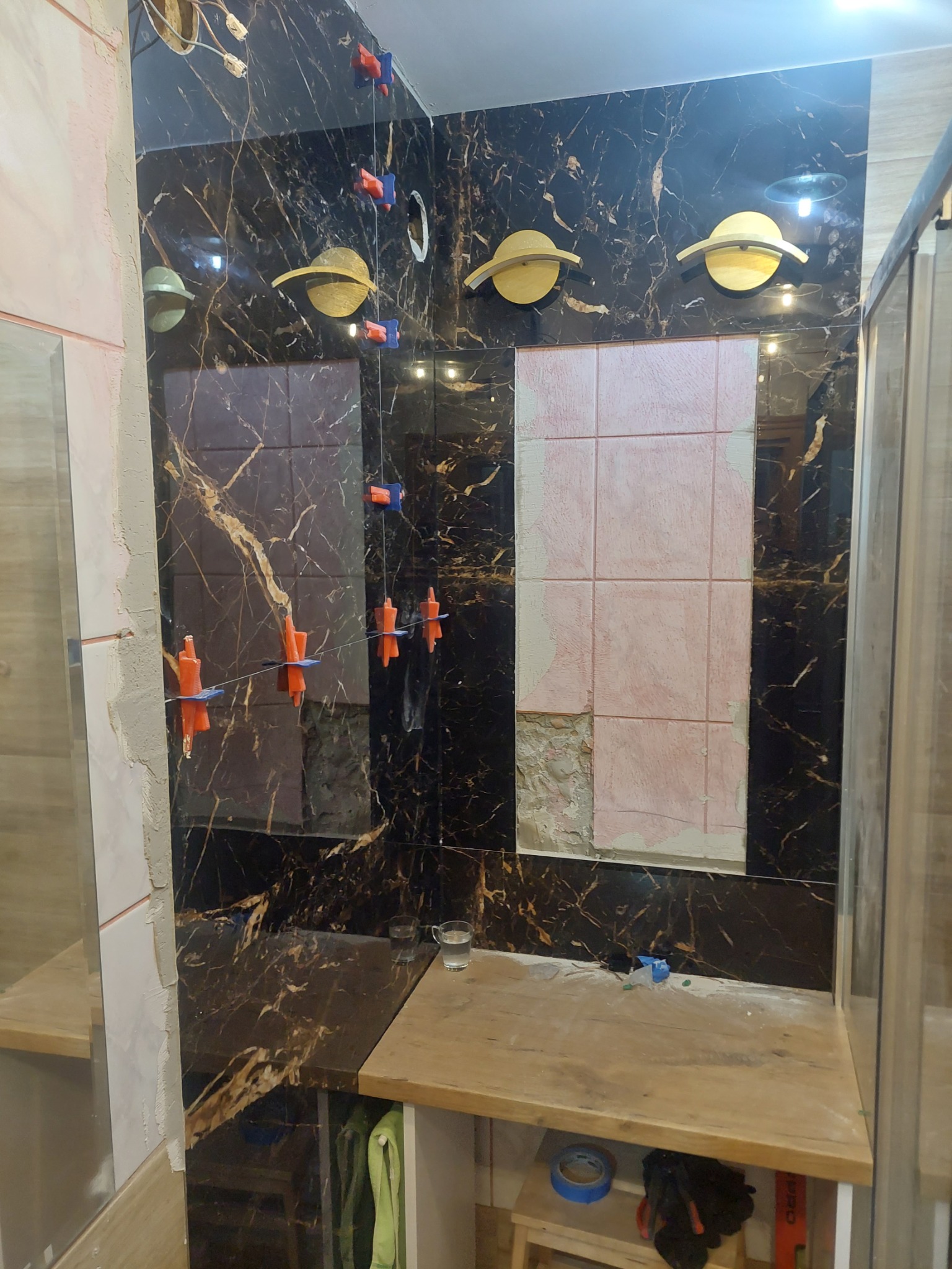 Montaż płytek imitujących marmur w łazience, widoczne systemy poziomowania płytek i fragment ściany przygotowanej pod płytki. Oświetlenie w stylu retro.