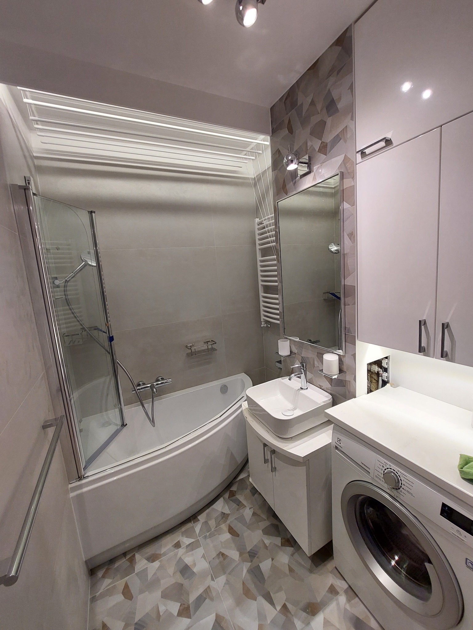 Nowoczesna łazienka po remoncie: narożna wanna z parawanem, umywalka nablatowa, pralka i szafki w bieli, mozaika na ścianie i podłodze. Oświetlenie punktowe.