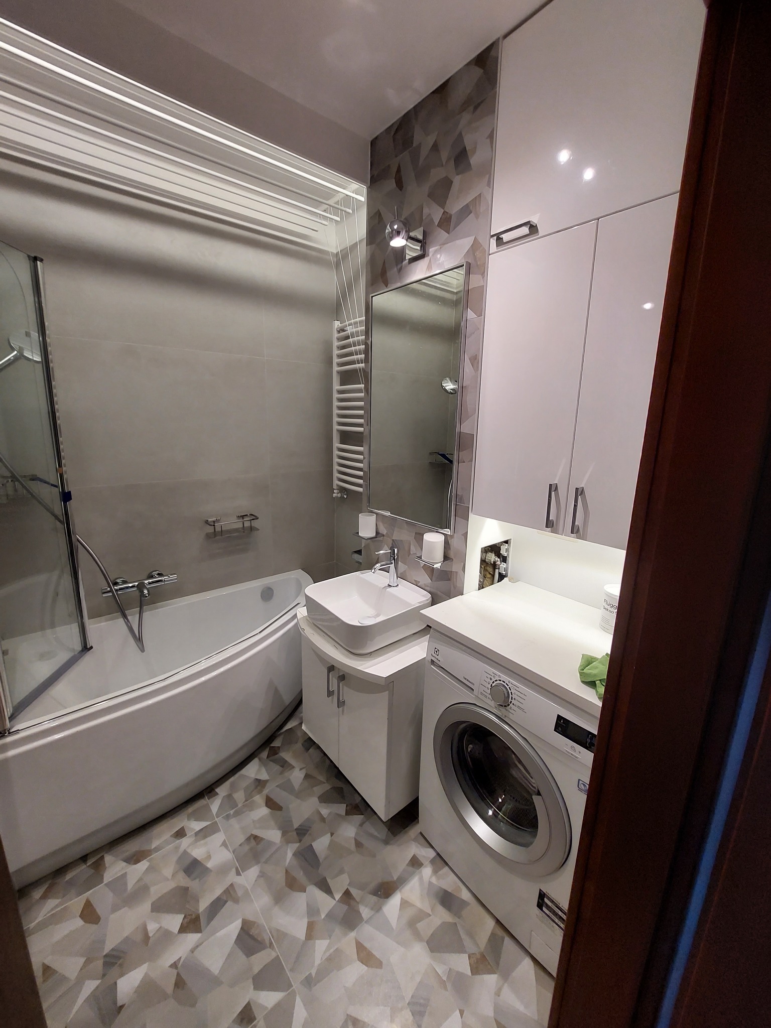 Nowoczesna łazienka z wanną, umywalką nablatową, pralką i geometryczną mozaiką na ścianie. Widoczna suszarka podsufitowa i oświetlenie punktowe.