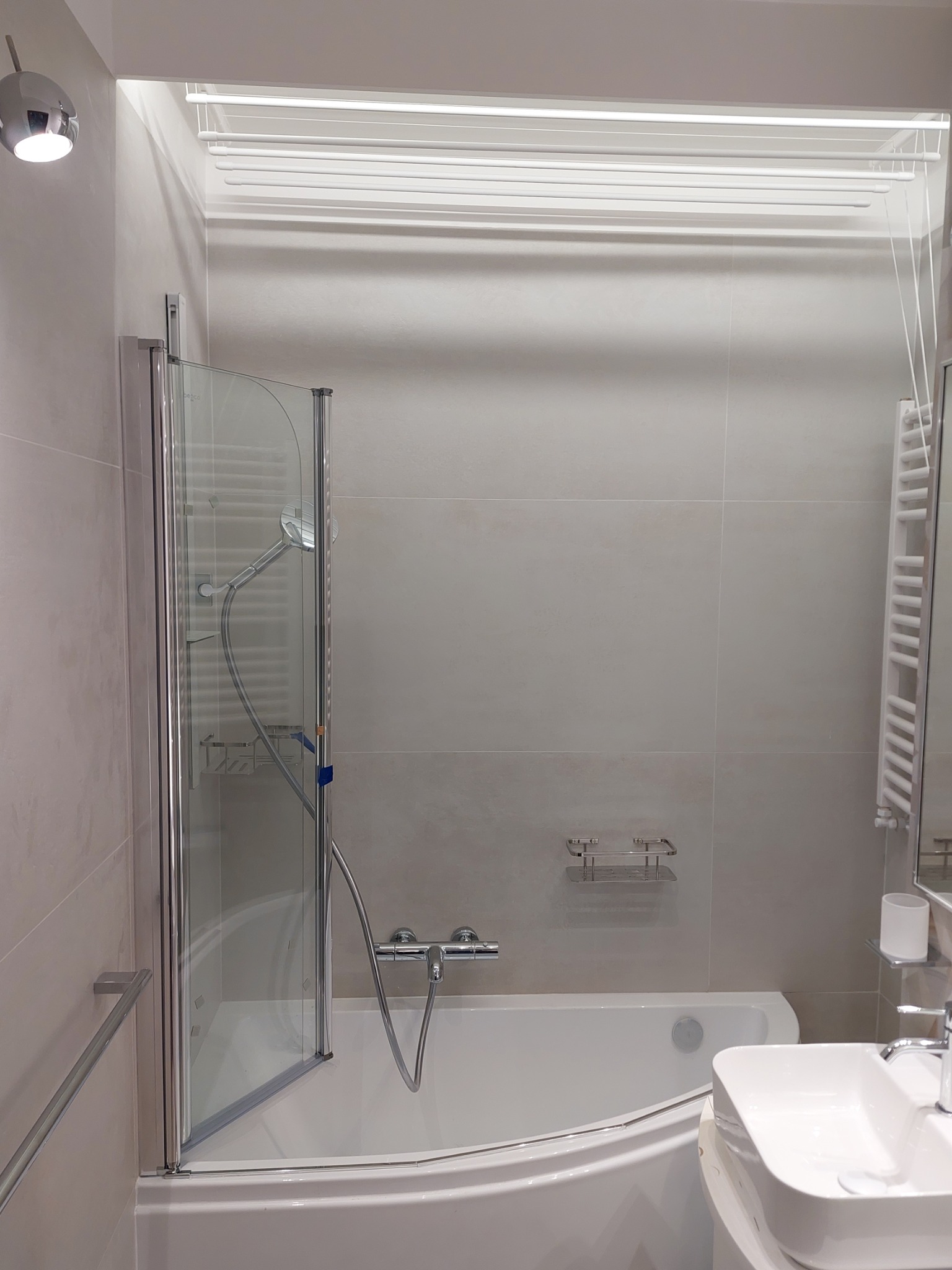 Jasna łazienka z wanną, szklanym parawanem nawannowym i umywalką. Na ścianach płytki w odcieniach beżu. Widoczna suszarka podsufitowa.