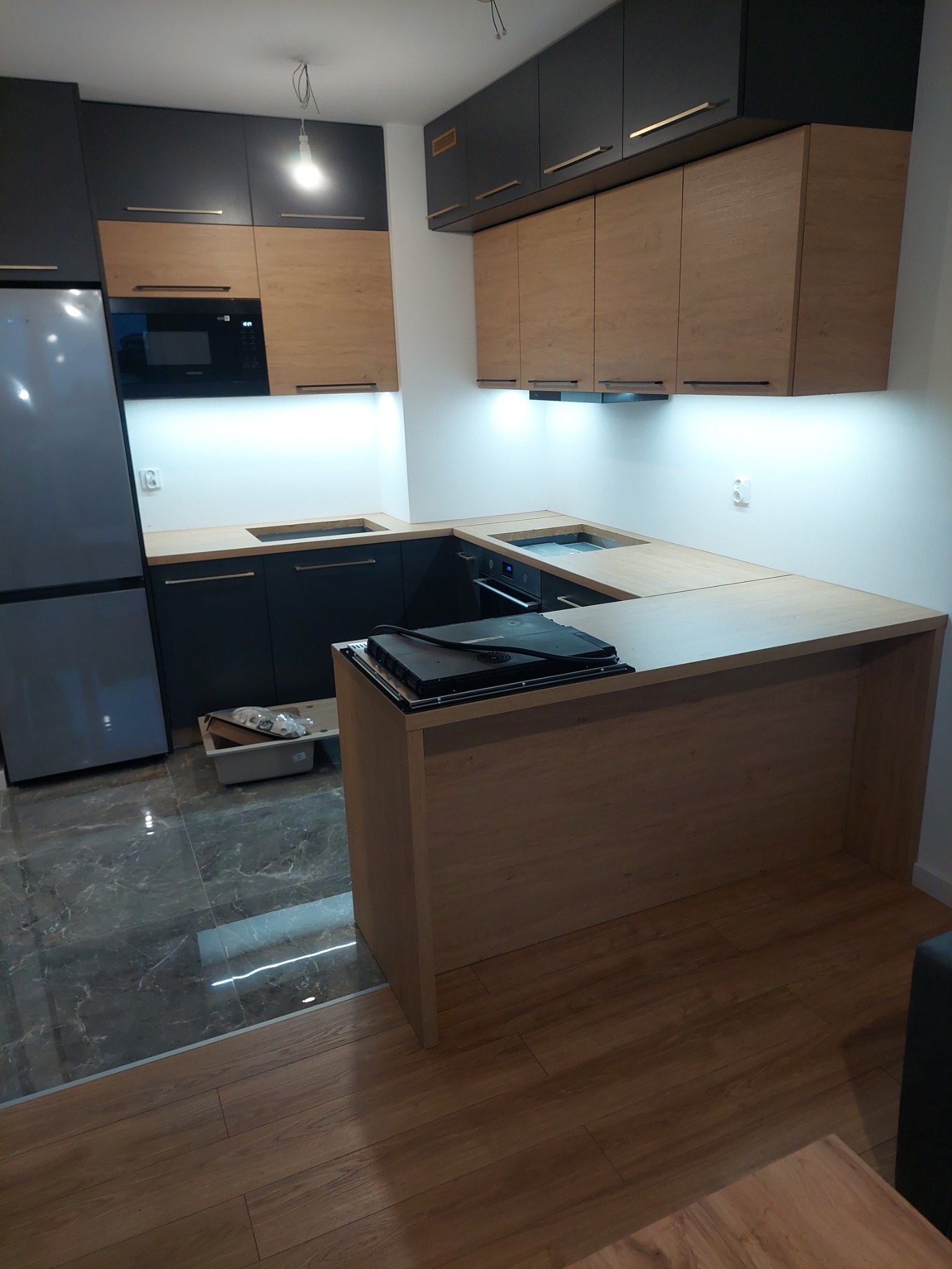 Nowoczesna kuchnia z czarnymi i drewnianymi szafkami, marmurową podłogą i drewnianym blatem wyspowym z płytą indukcyjną. Oświetlenie LED pod szafkami.