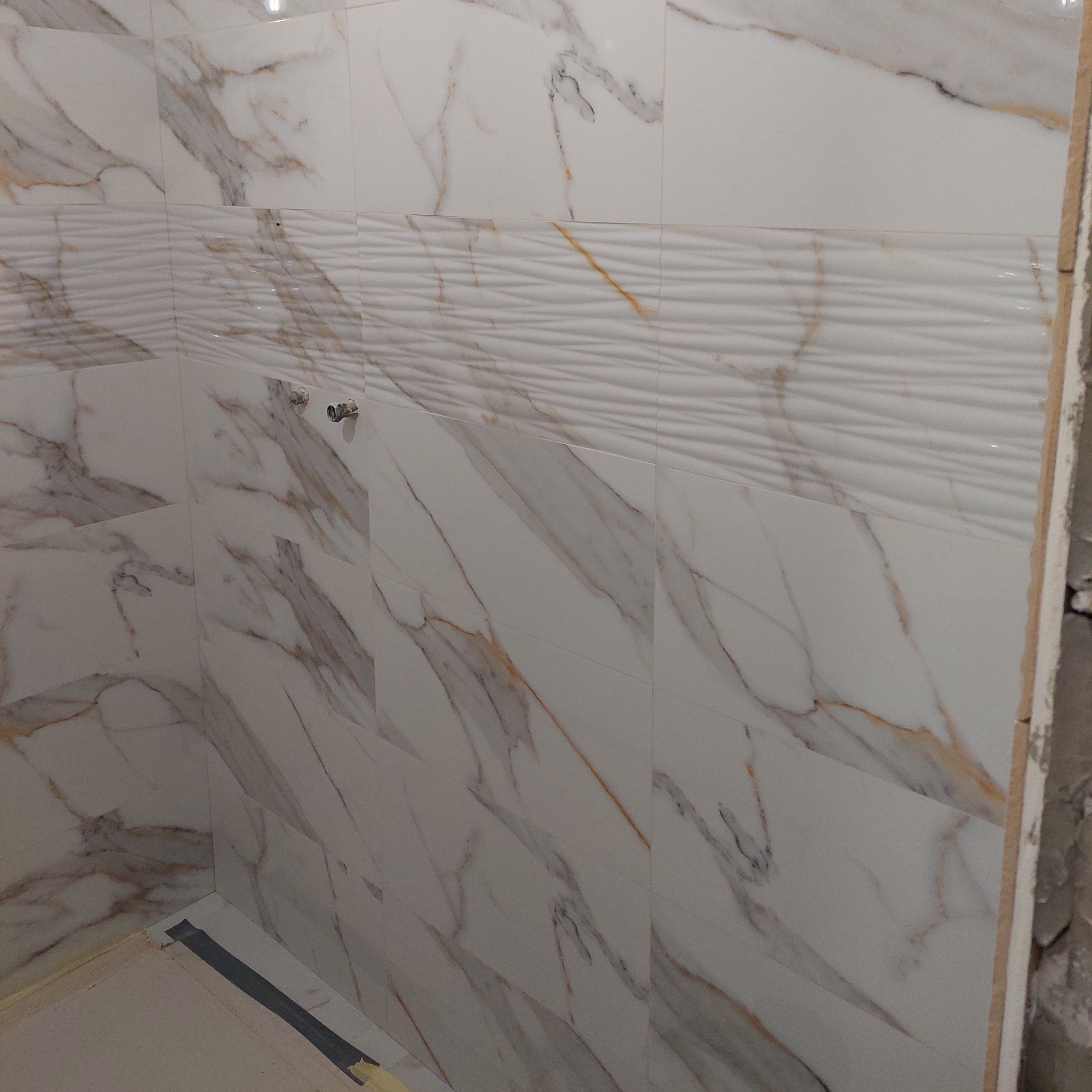 Ściana łazienki wyłożona białymi płytkami imitującymi marmur z poziomym pasem płytek o falistej strukturze. Widoczne fragmenty instalacji wodnej i surowa krawędź ściany.