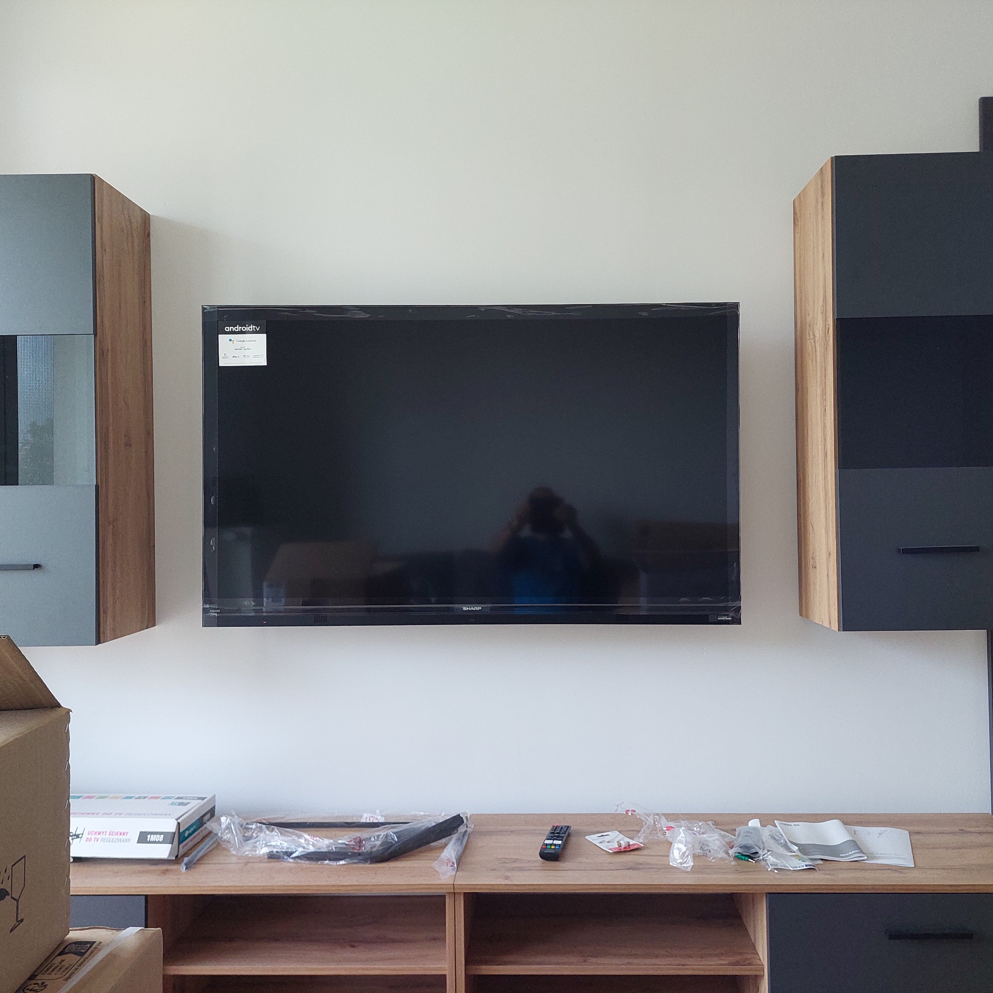 Telewizor Sharp Android TV zamontowany na ścianie między szafkami. Na szafce pod spodem akcesoria montażowe i pilot. Widoczny karton po uchwycie ściennym.