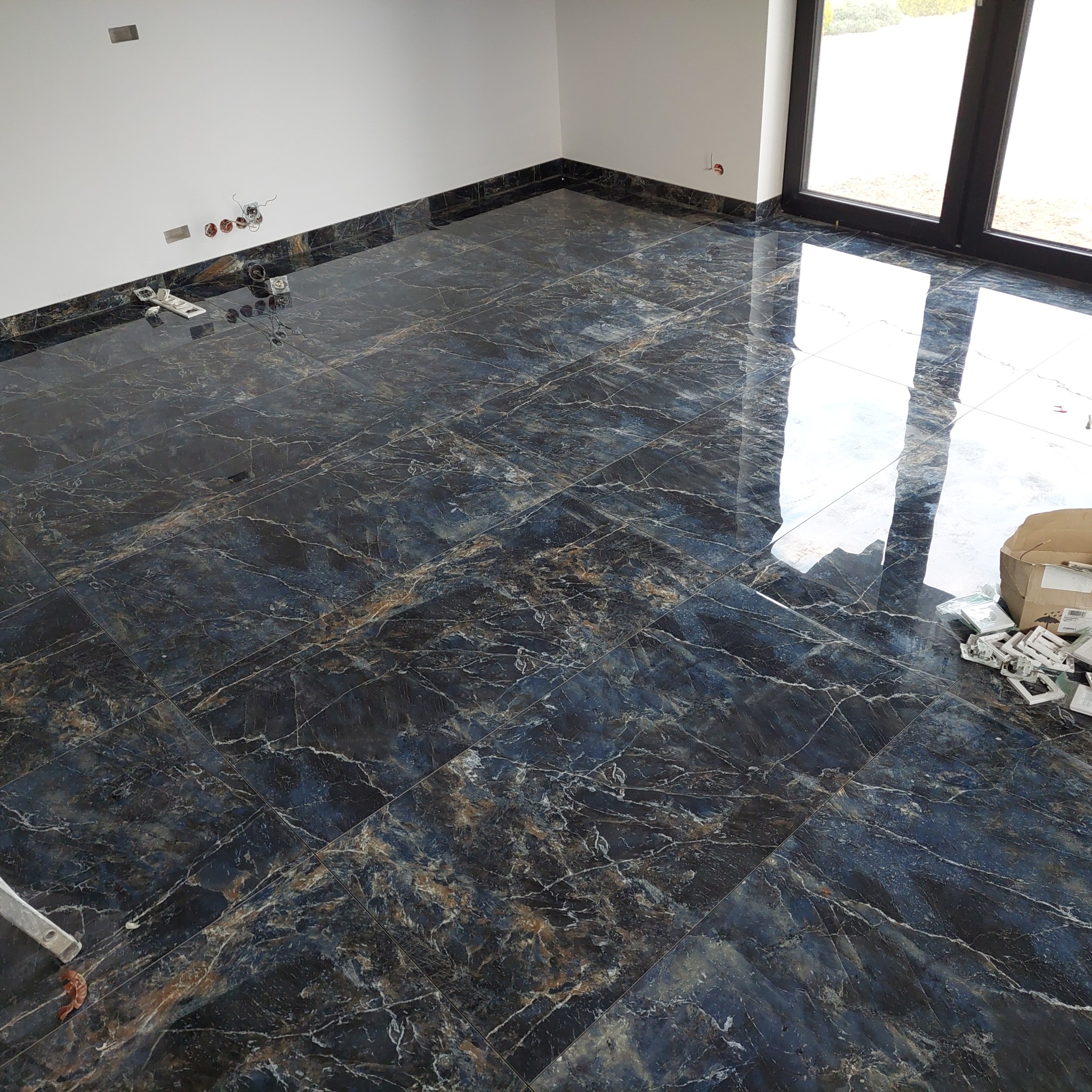 Położona, błyszcząca, ciemna podłoga z płytek imitujących marmur w nowym wnętrzu. Widoczne puszki elektryczne i listwy przypodłogowe z tego samego materiału.