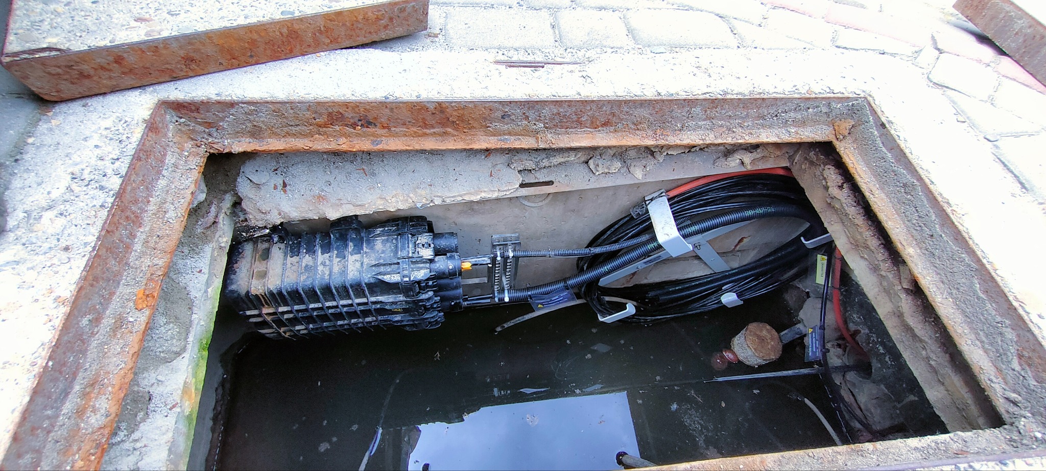 Studzienka telekomunikacyjna z kablem światłowodowym i czarną puszką łączeniową, widok z góry, zardzewiała rama włazu, woda na dnie.